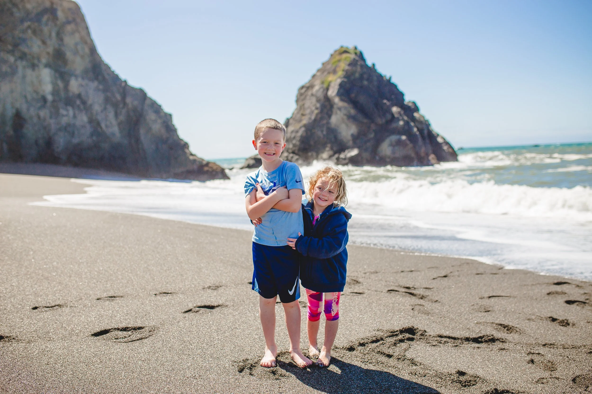 Two children, a boy and a girl, standing arm-in-arm on a sandy beach with large rock formations and the ocean in the background. The boy is crossing his arms and smiling, wearing a blue t-shirt and dark shorts. The girl is smiling, wearing a navy hoo