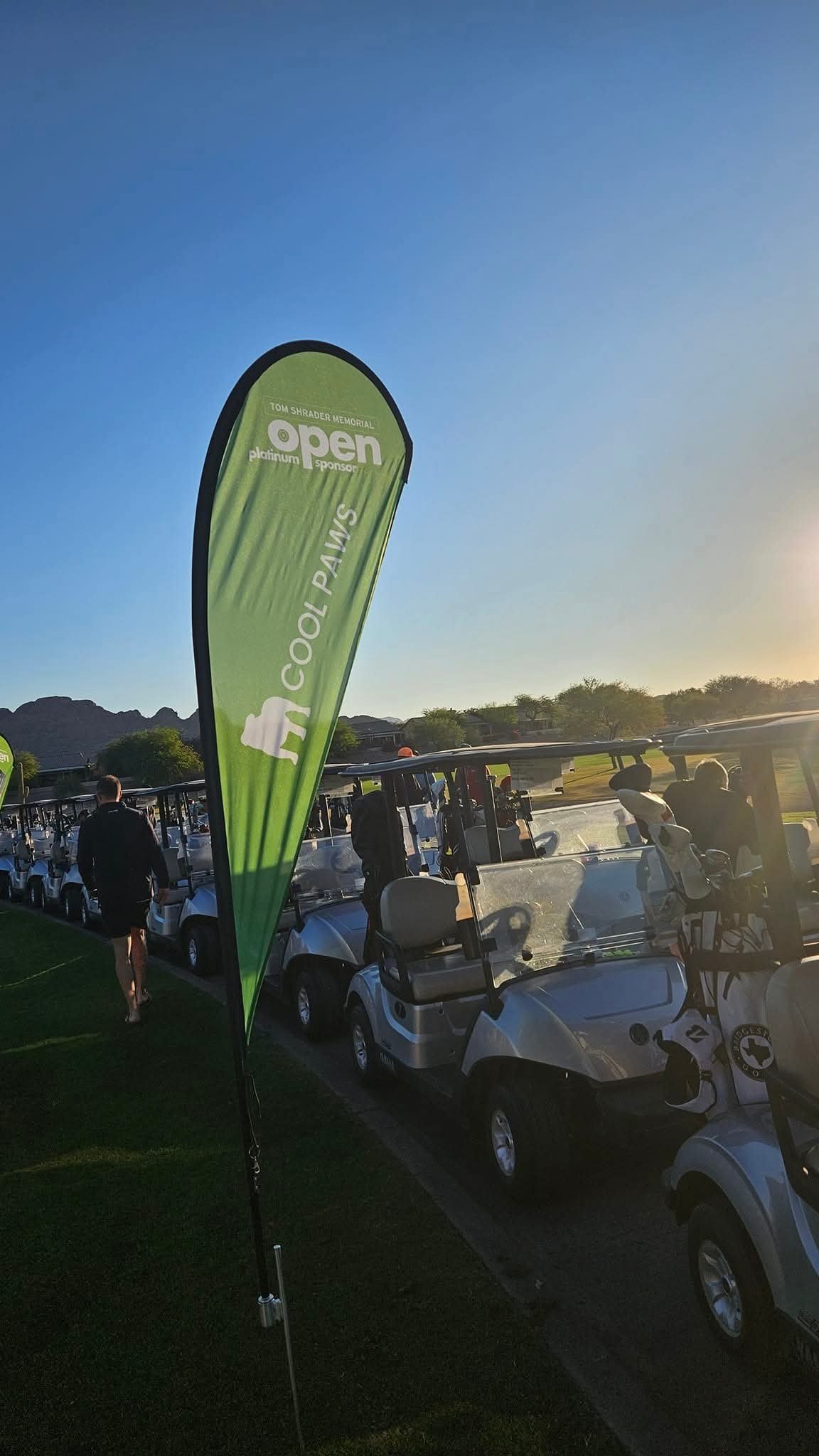 Golf carts lined up at a golf course with a green banner displaying 'Tom Shrader Memorial Open Platinum Sponsor' and 'Cool Pains' logo, under a clear sky with the sun setting.