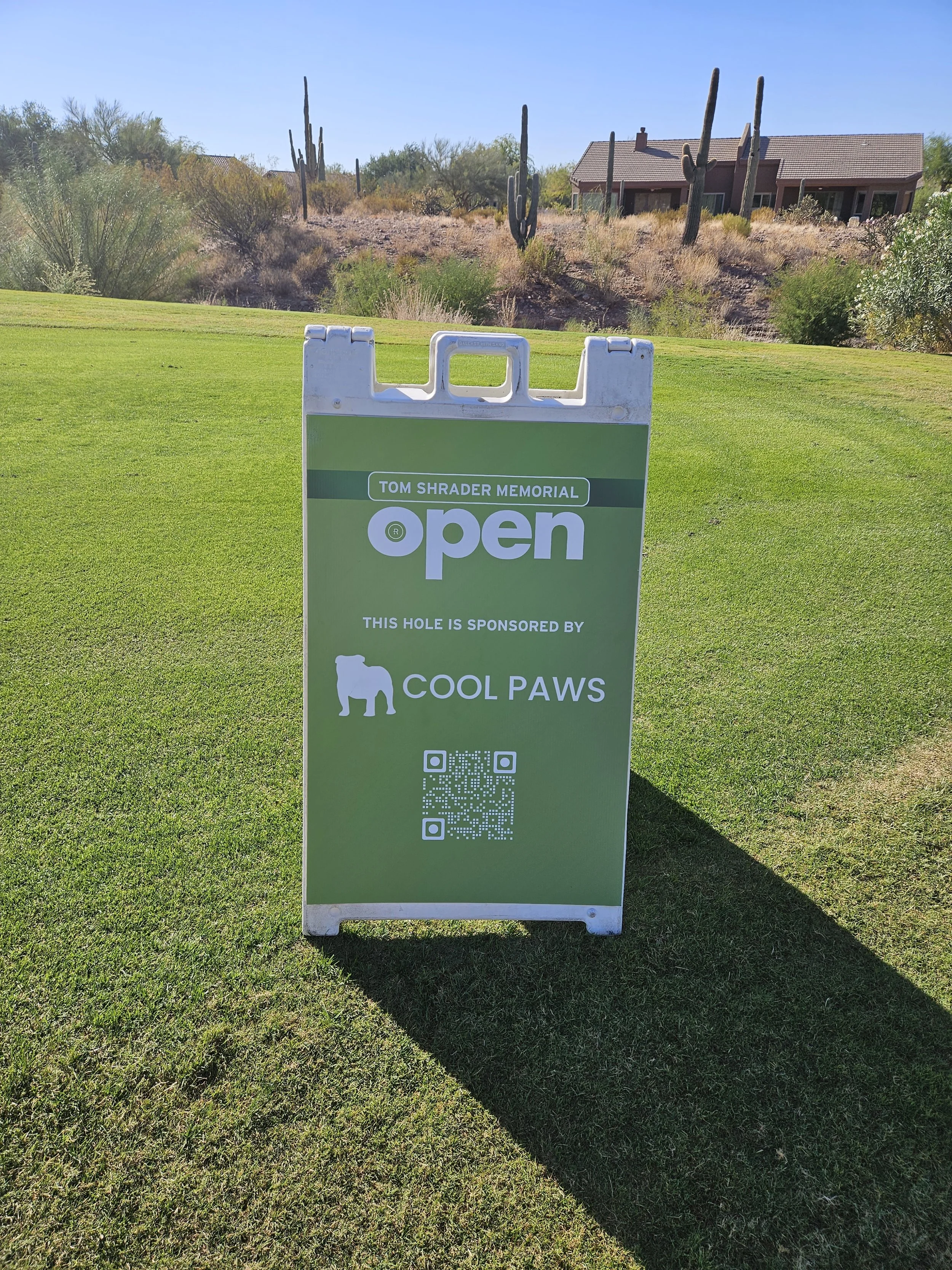 Sign on a golf course advertising the Tom Shrader Memorial open, sponsored by Cool Paws, with a QR code at the bottom.