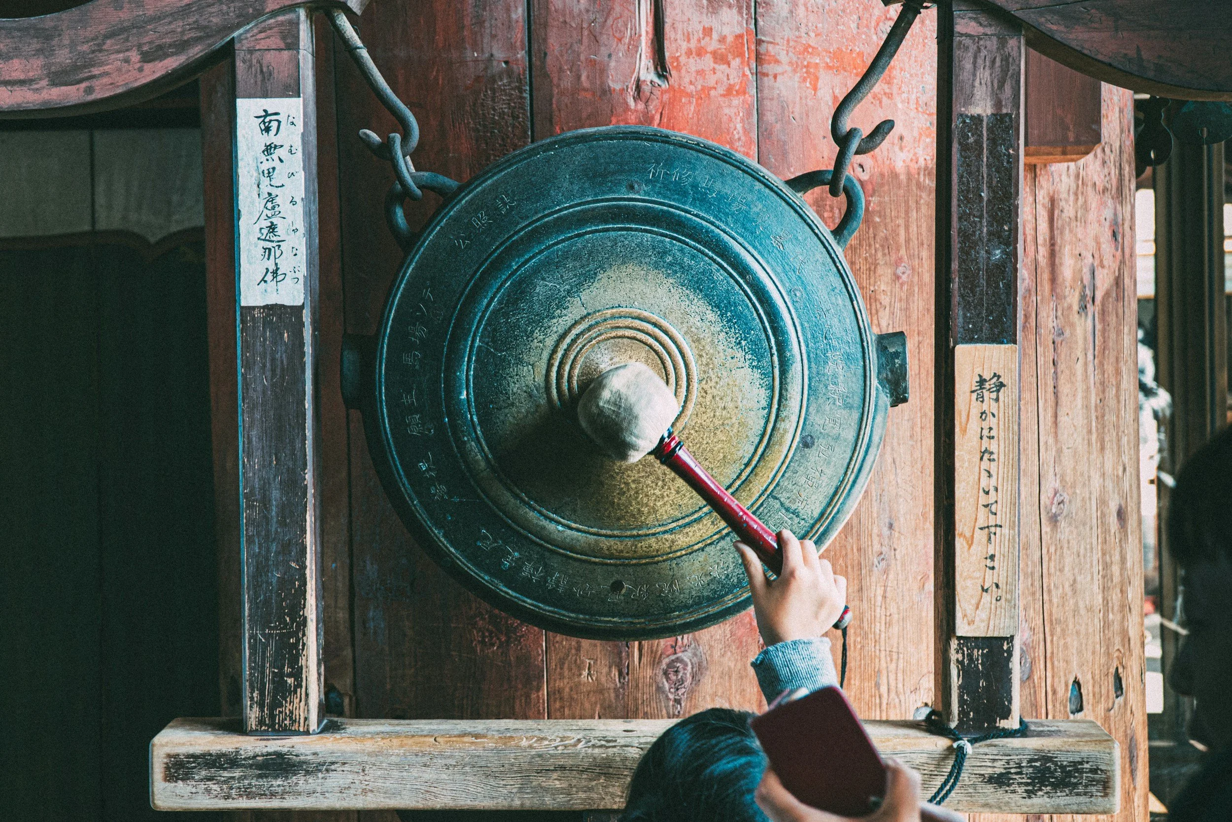 A large gong mounted on a wooden structure being struck with a mallet by a person in a gray sleeve, with two other people nearby observing.