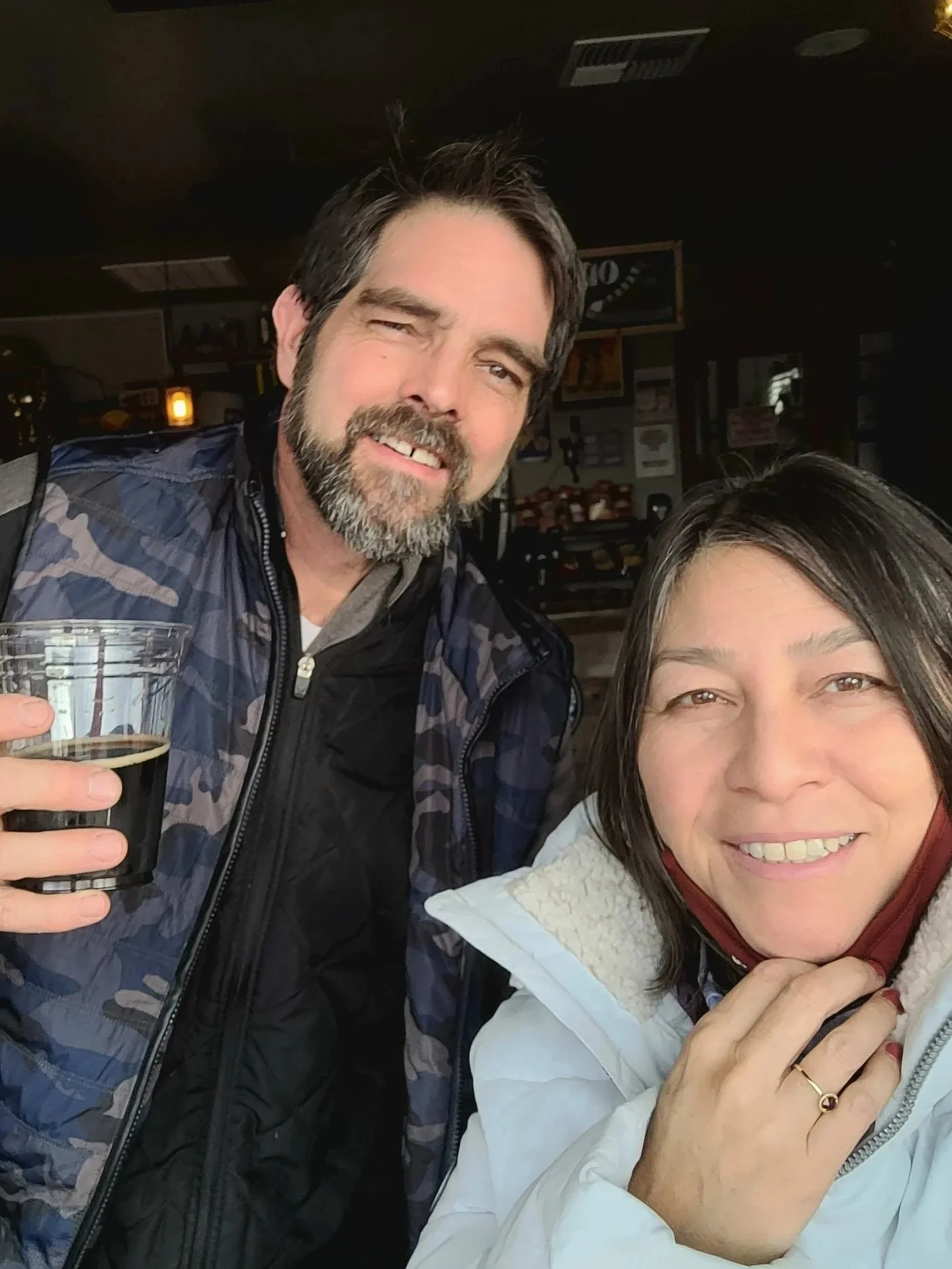A man and woman smiling indoors, with the man holding a glass of dark beverage. The woman has a red mask pulled down under her chin, and they are close together.