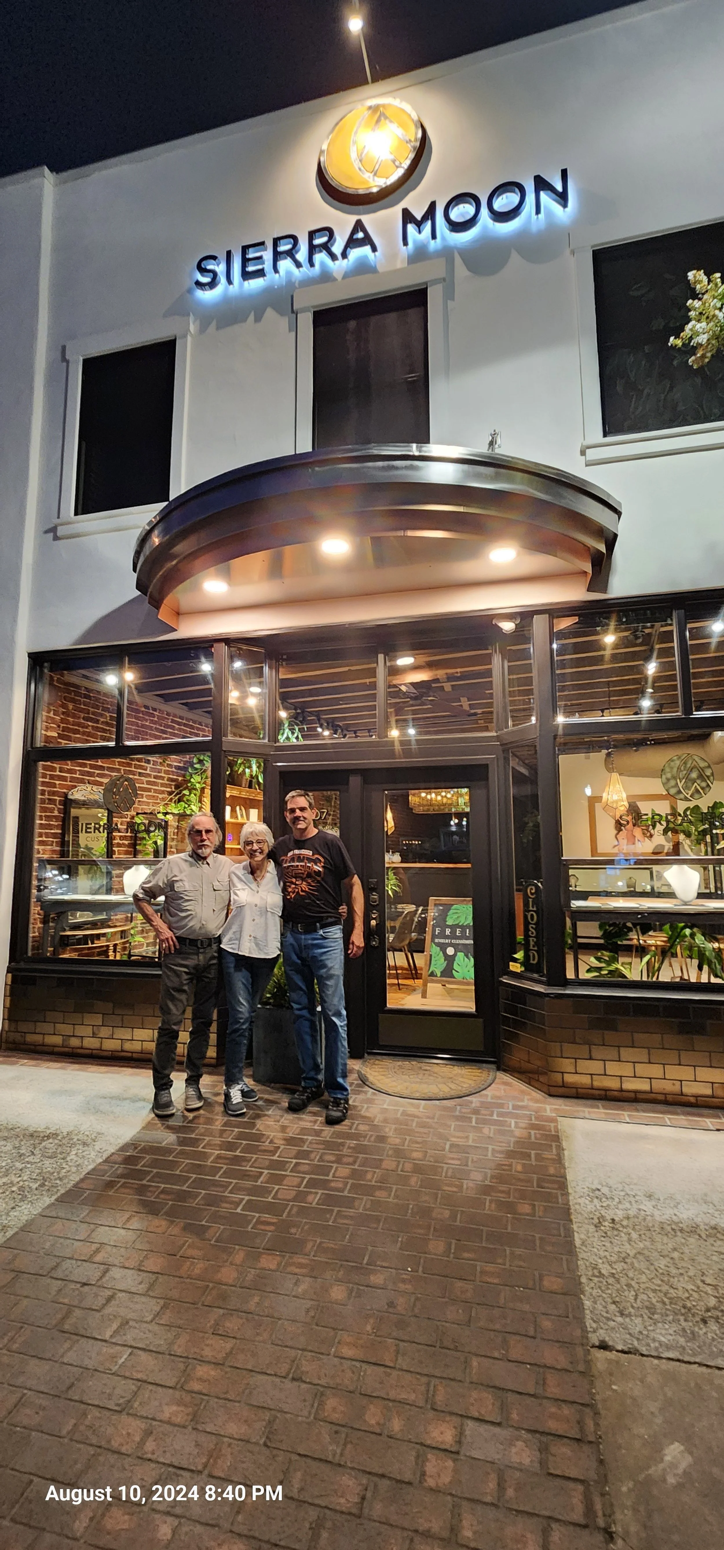 Nighttime photo of Sierra Moon restaurant with three people standing in front, smiling. The restaurant has large glass windows, a brick exterior, and a neon sign. The date and time shown are August 10, 2024, at 8:40 PM.