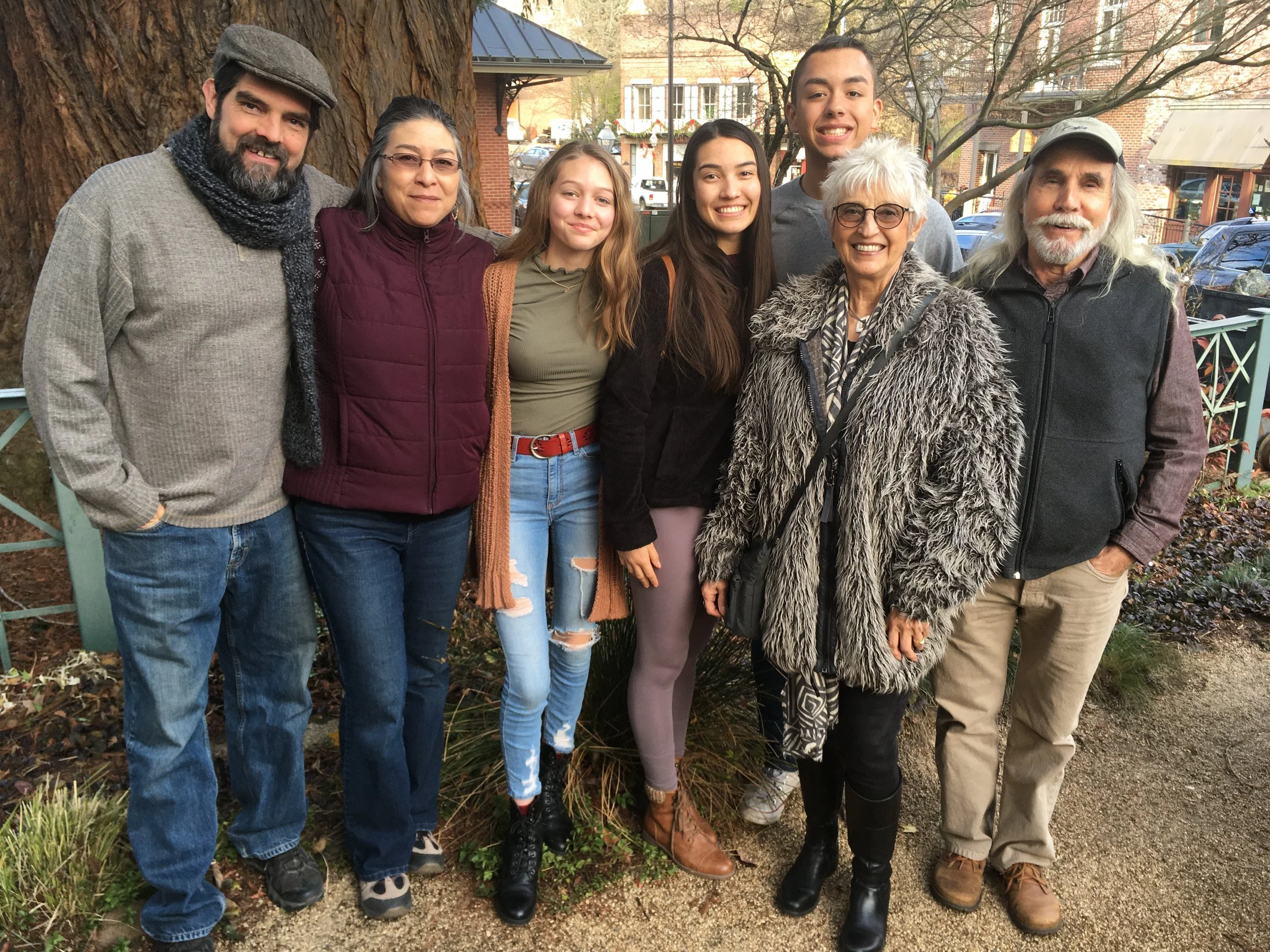 Group of seven people standing outdoors in front of a large tree, smiling for the photo, with autumn trees and buildings in the background.