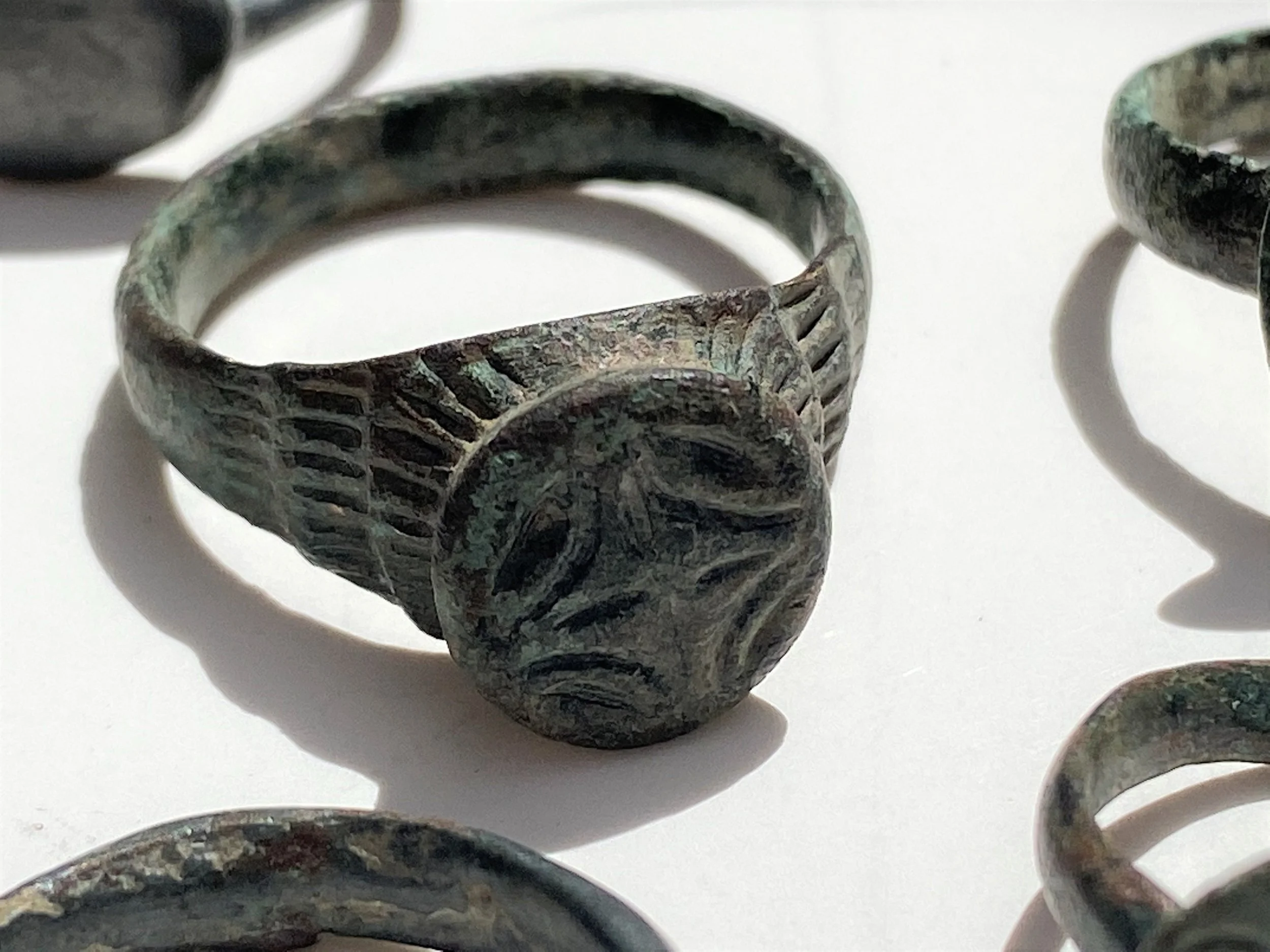 Close-up of an ancient ring with a carved face, made of a weathered material, displayed with other similar rings on a white surface.