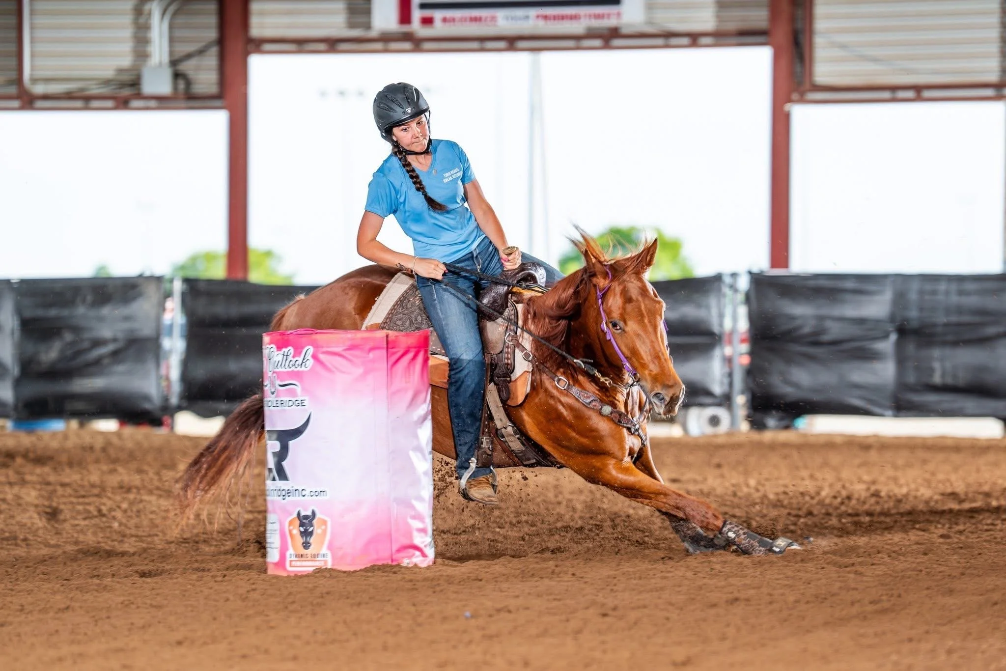 A woman riding in rodeo royalty gear on a horse in an indoor equestrian arena, navigating around a pink barrel with a logo, as part of a barrel racing event. Rodeo Royalty