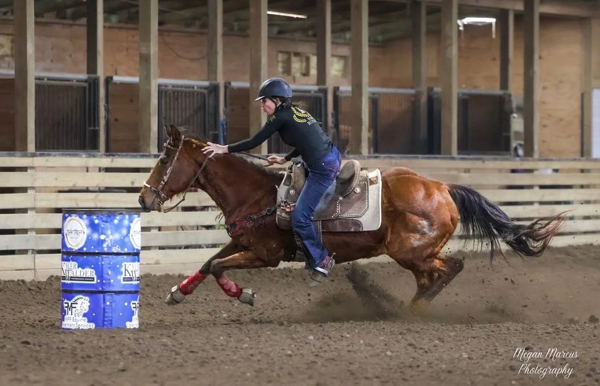 A person riding a horse in an indoor equestrian arena, navigating around a blue barrel during a barrel racing event wearing rodeo royalty gear.