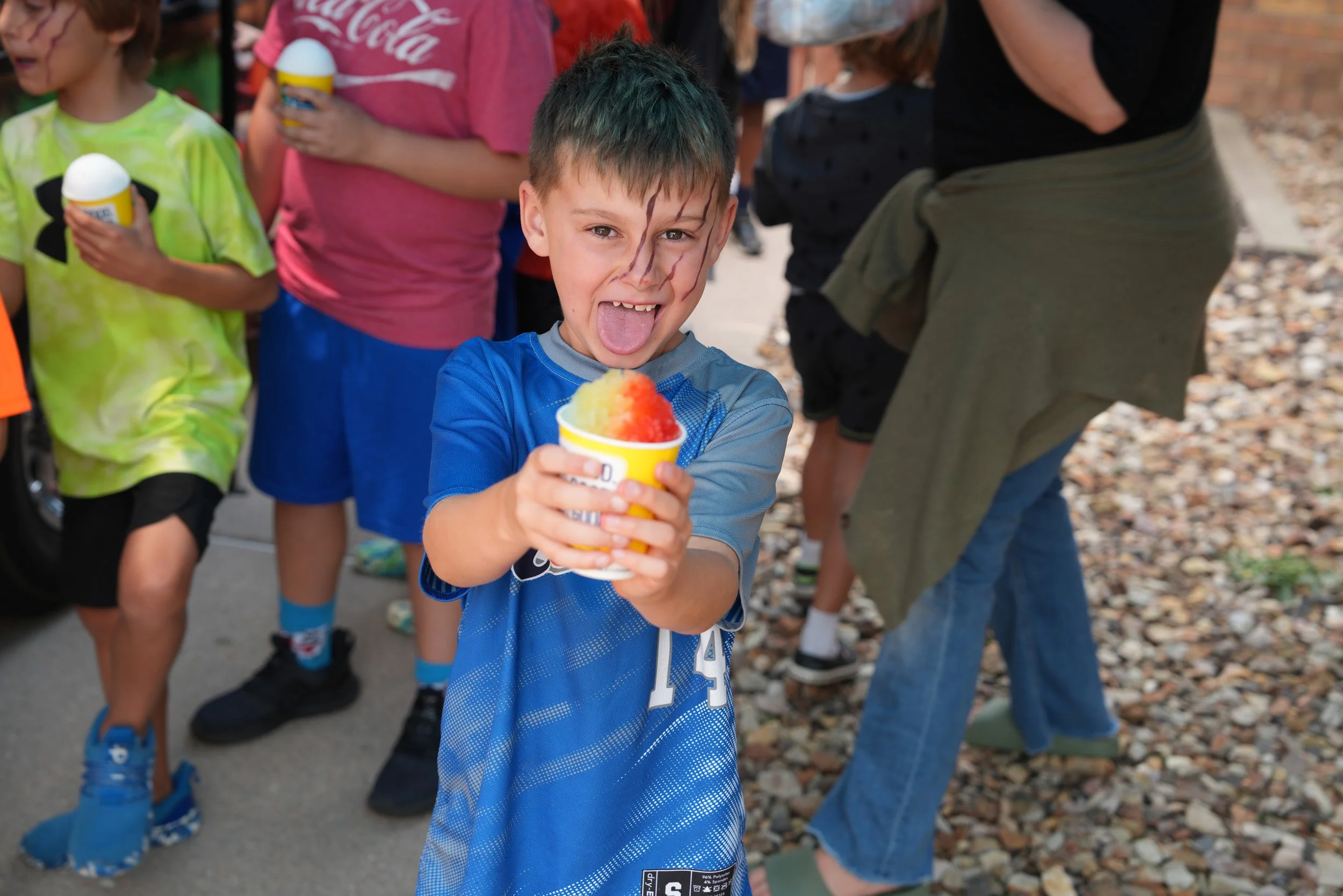 A young boy with face paint on his forehead and cheeks, holding a cup of shaved ice, sticking out his tongue, and smiling at the camera. Other children and adults hold similar cups in the background during an outdoor event.