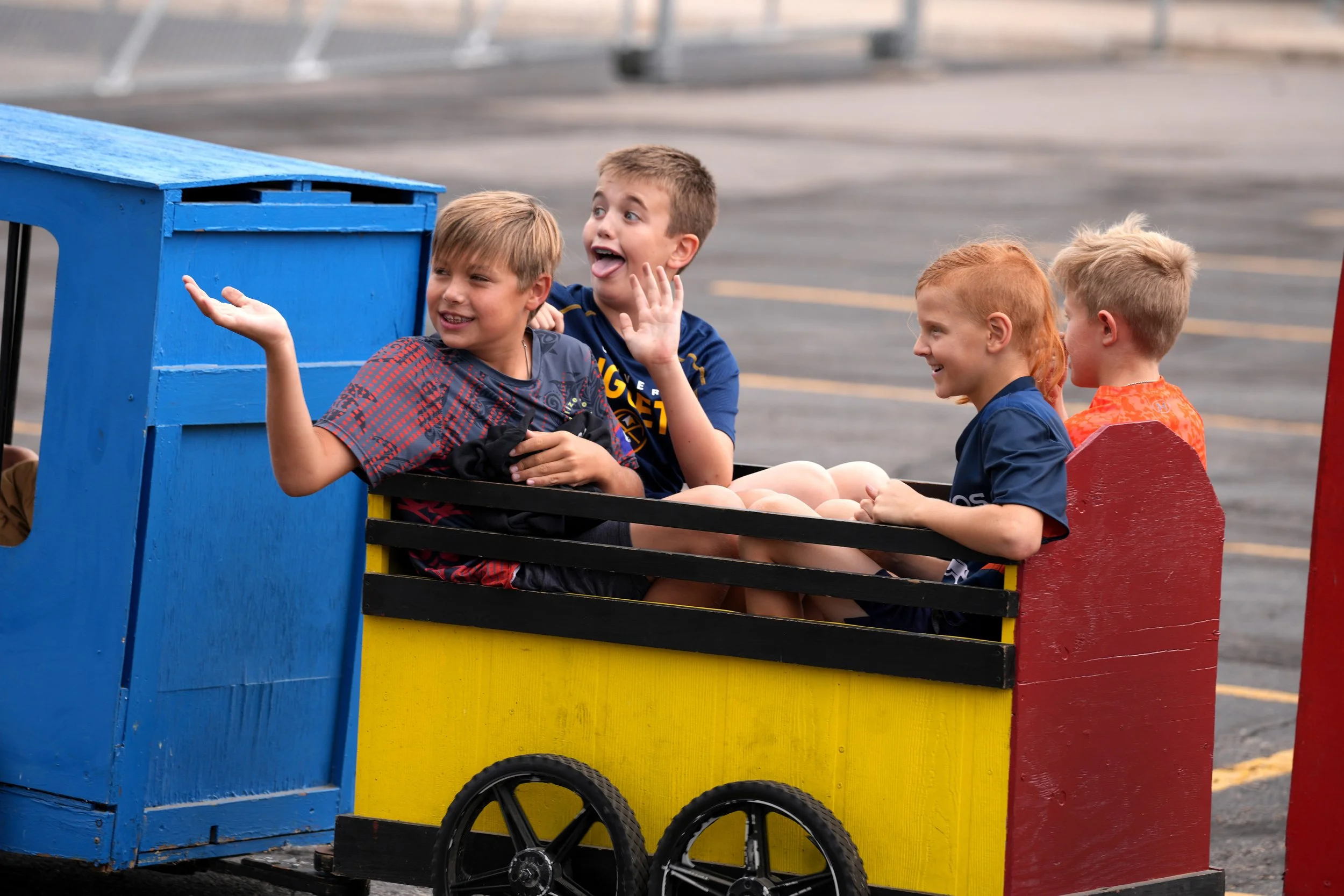 Children riding an amusement park train painted in blue yellow and red, with a black wheel visible, on a paved surface.