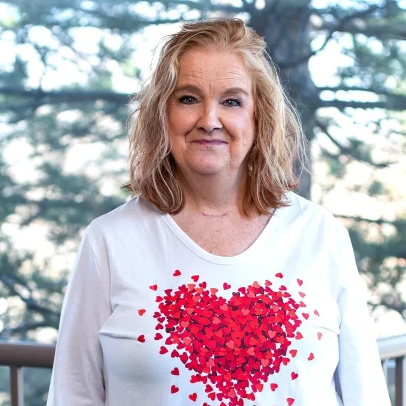 A woman with blonde, wavy hair wearing a white shirt with a heart made of small red hearts printed on it, standing outdoors with trees in the background.