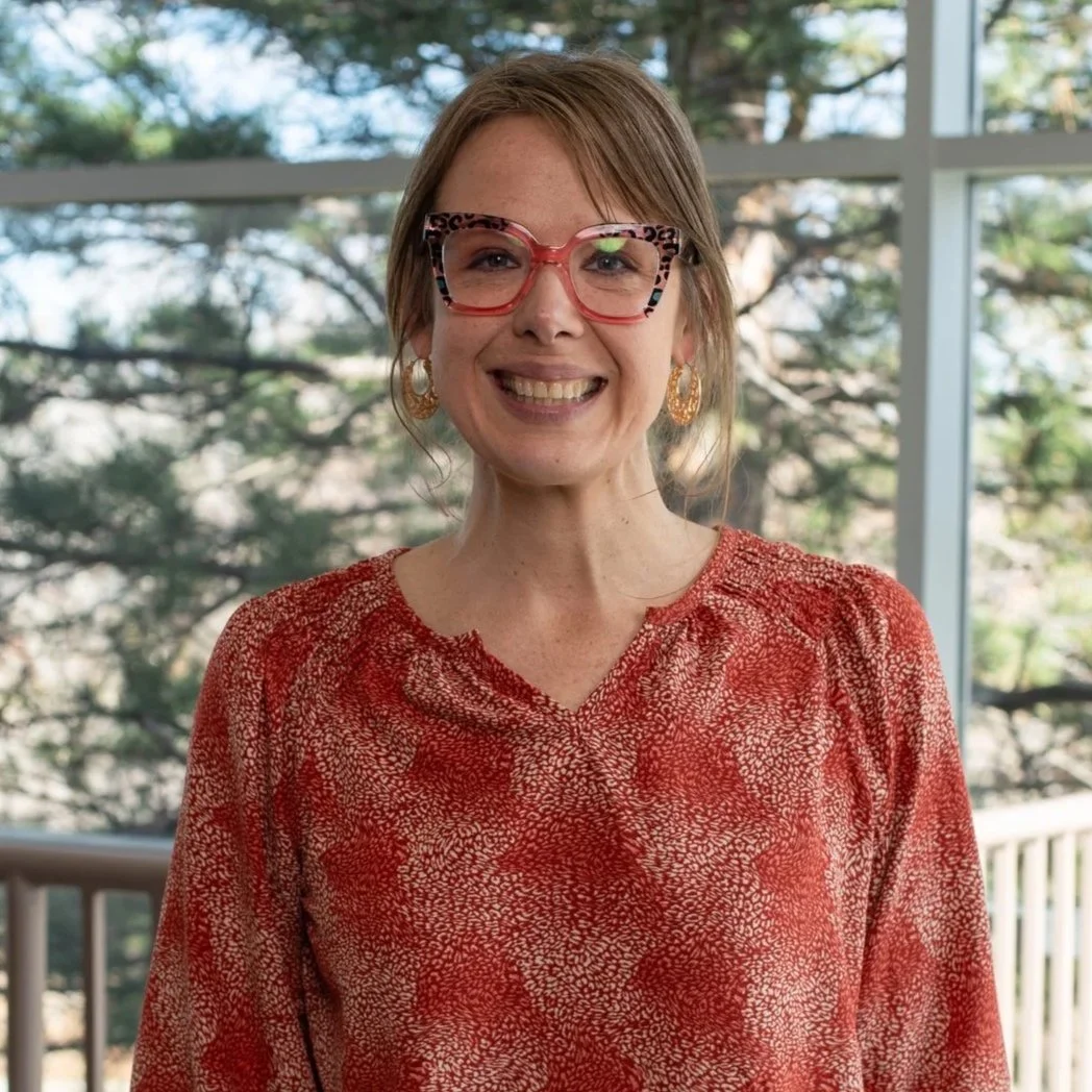 A woman with short hair wearing red glasses, gold earrings, and a red patterned top, smiling in front of a window with trees outside.