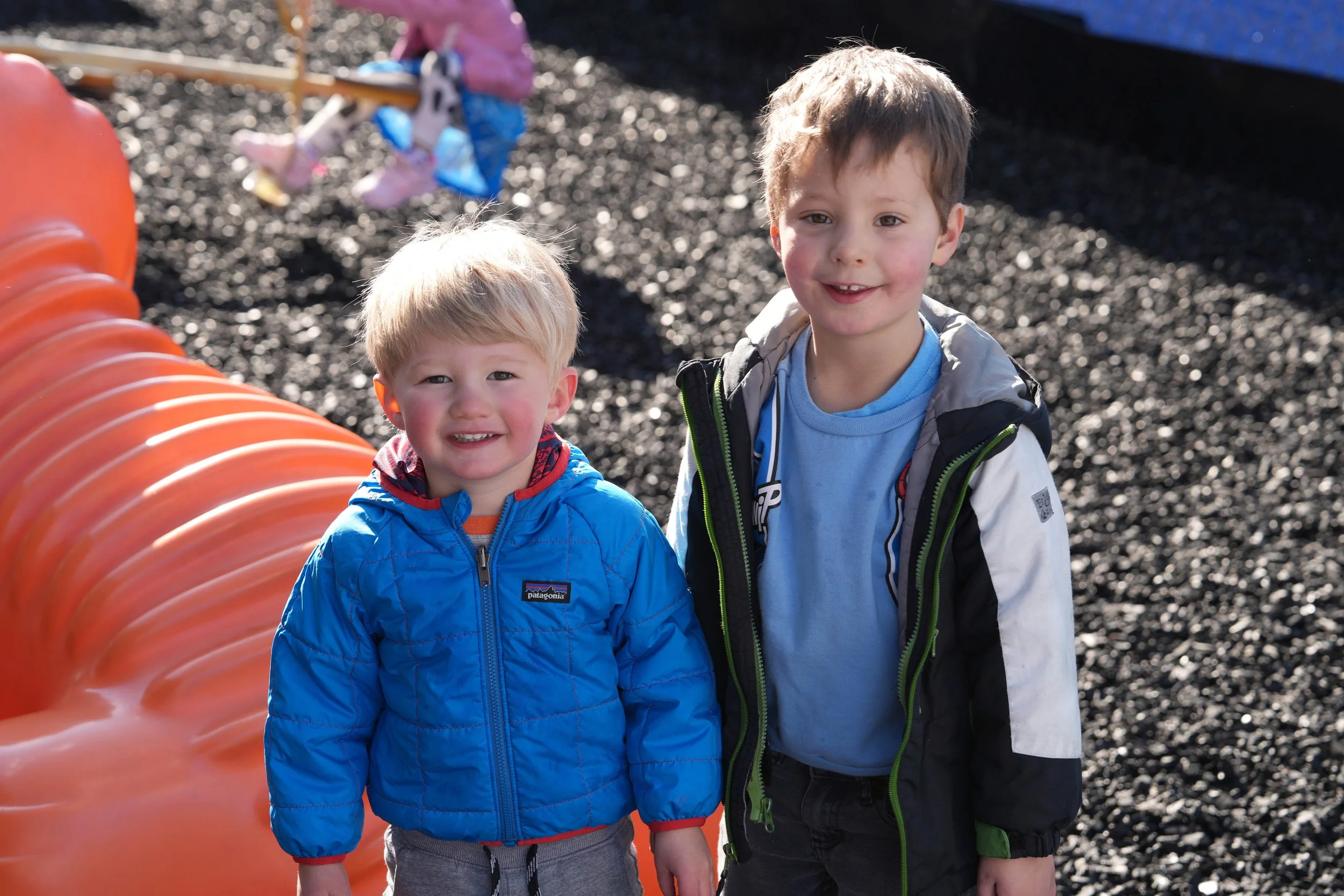 Two young boys standing outdoors next to an orange playground tunnel on a surface covered with black gravel. The boy on the left has blonde hair, is wearing a bright blue jacket, and is smiling at the camera. The boy on the right has brown hair, is wearing a light blue shirt and a beige and green jacket, and is also smiling at the camera. In the background, a girl is playing on a swing set.