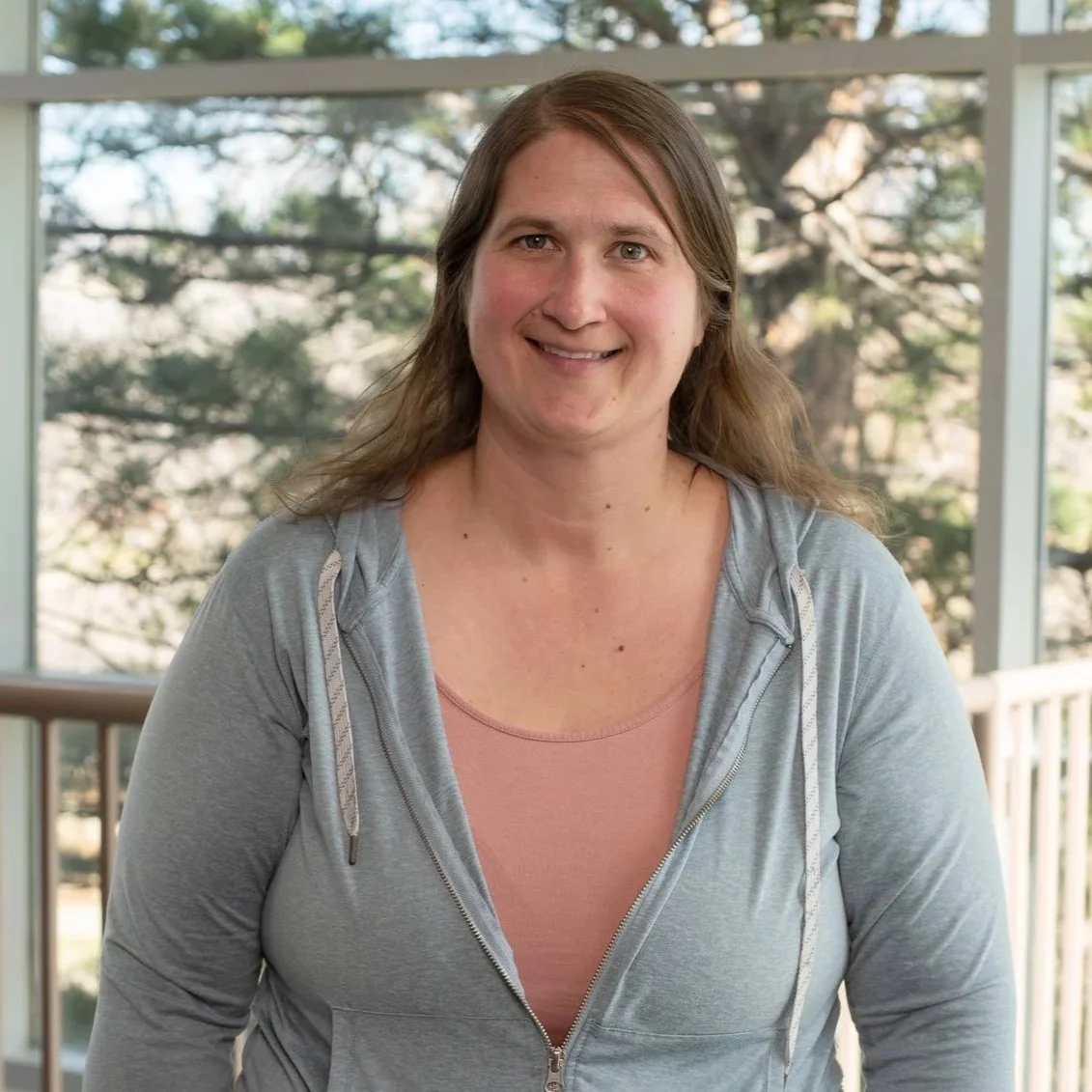 A woman with brown hair and a light gray hoodie, smiling, standing outside with trees and a porch railing in the background.