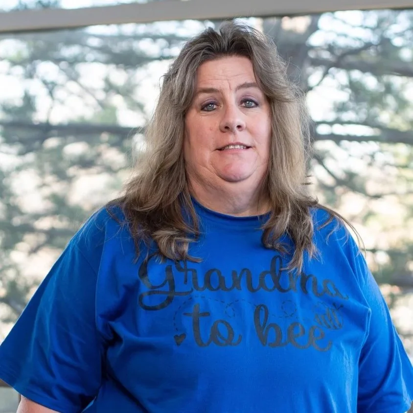 A woman with long, wavy hair wearing a blue T-shirt that says 'Grandma to bee',