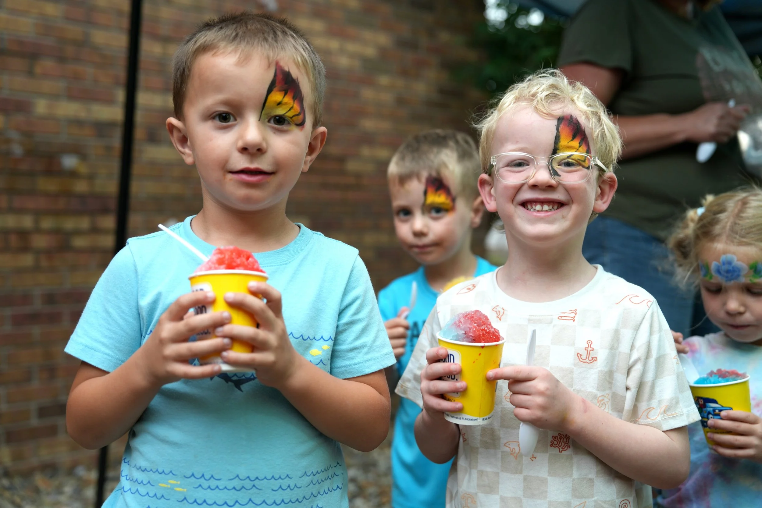 A group of children holding colorful snow cones at an outdoor gathering, with face paint on their faces and a brick wall in the background.