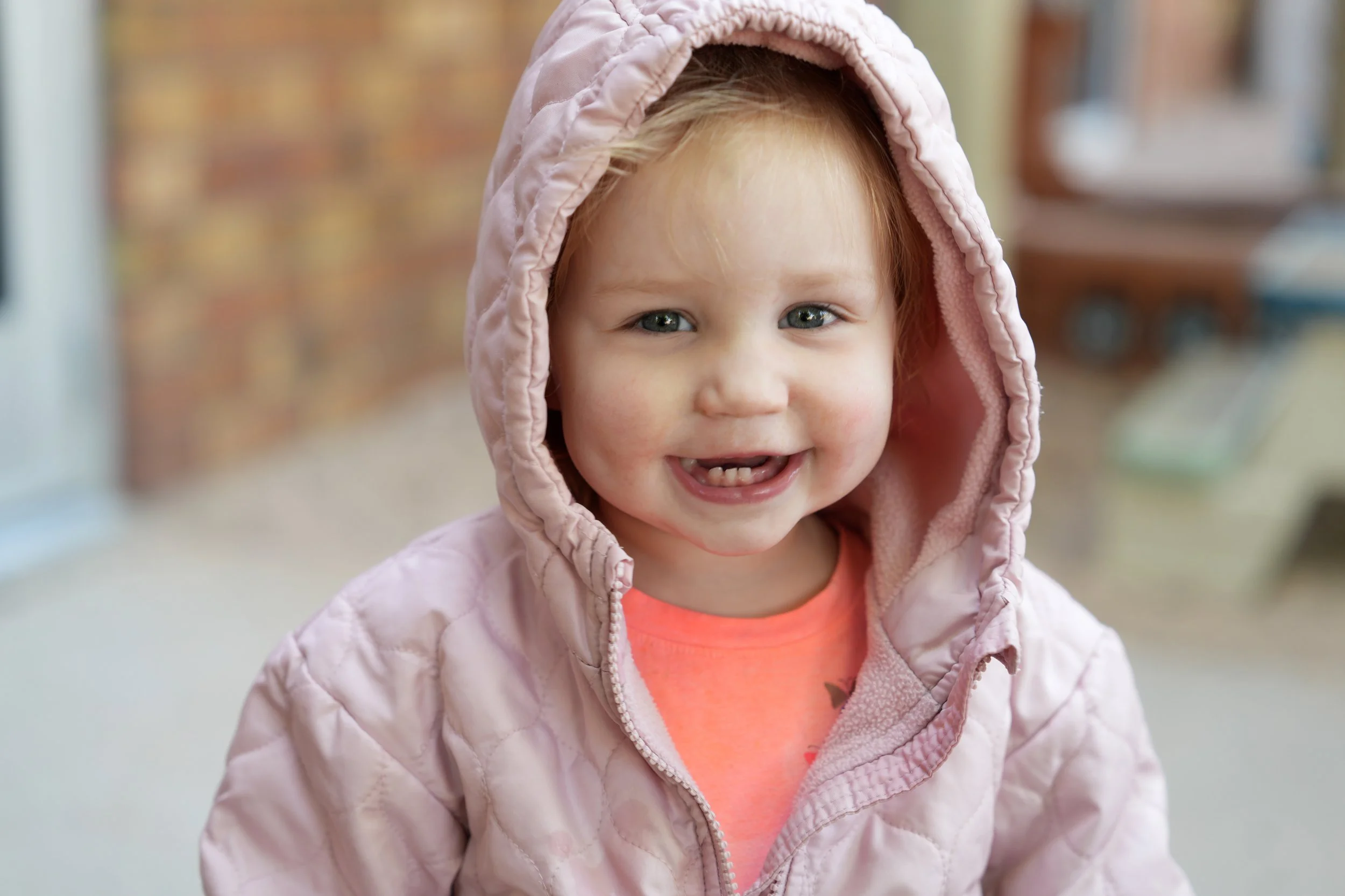 A young girl with red hair, blue eyes, and a big smile, wearing a light pink quilted hooded jacket and an orange shirt, standing outdoors with a blurred background.