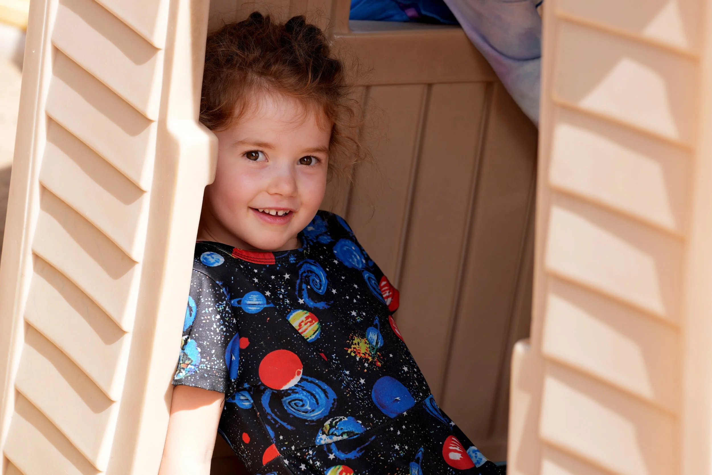 A young girl with curly hair and space-themed clothing peeks out from behind beige plastic shutters, smiling.