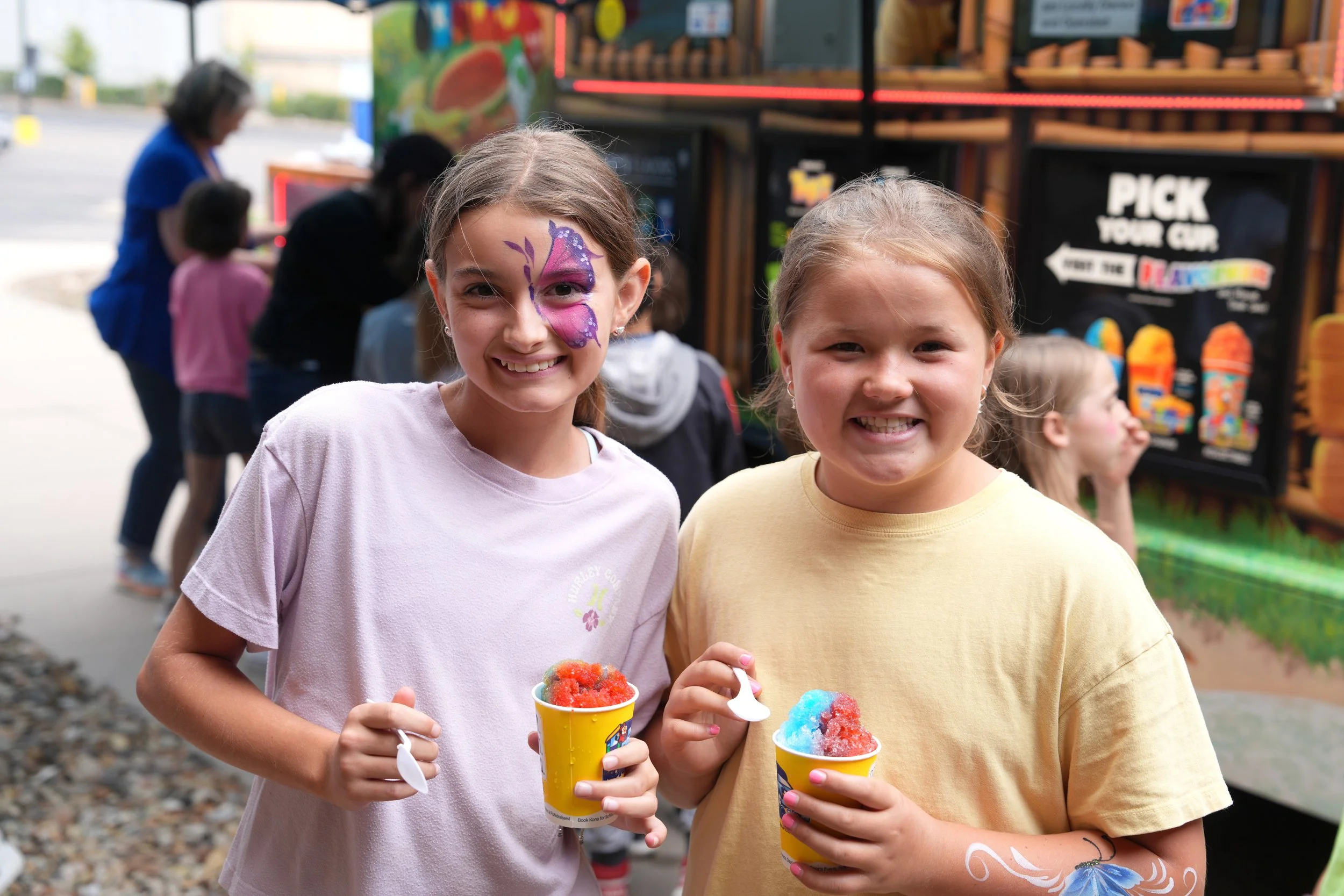 Two young girls smiling and holding colorful snow cones at an outdoor fair or festival, with a food truck and other people in the background.