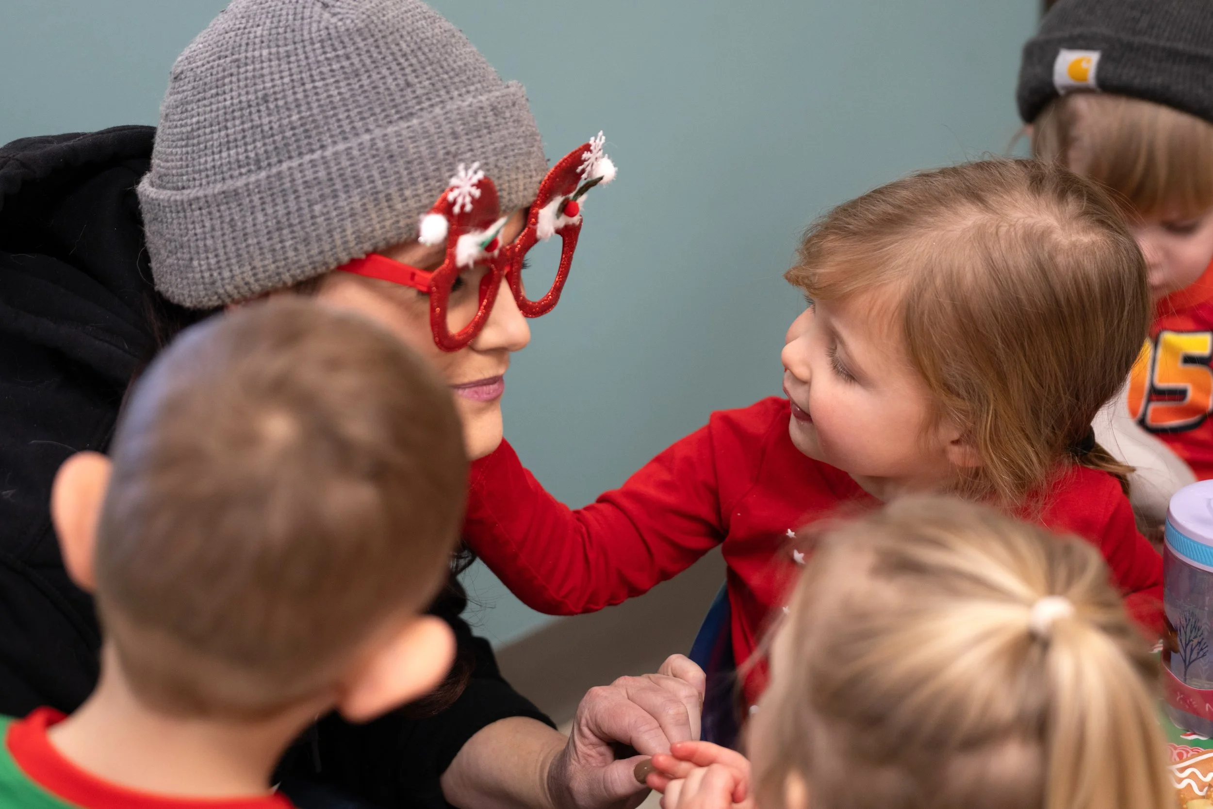 A woman wearing novelty red glasses with Christmas decorations, a gray beanie, and a black jacket interacting with children. The children are wearing colorful clothing and hats, and are gathered around the woman, engaging with her in a festive gathering.