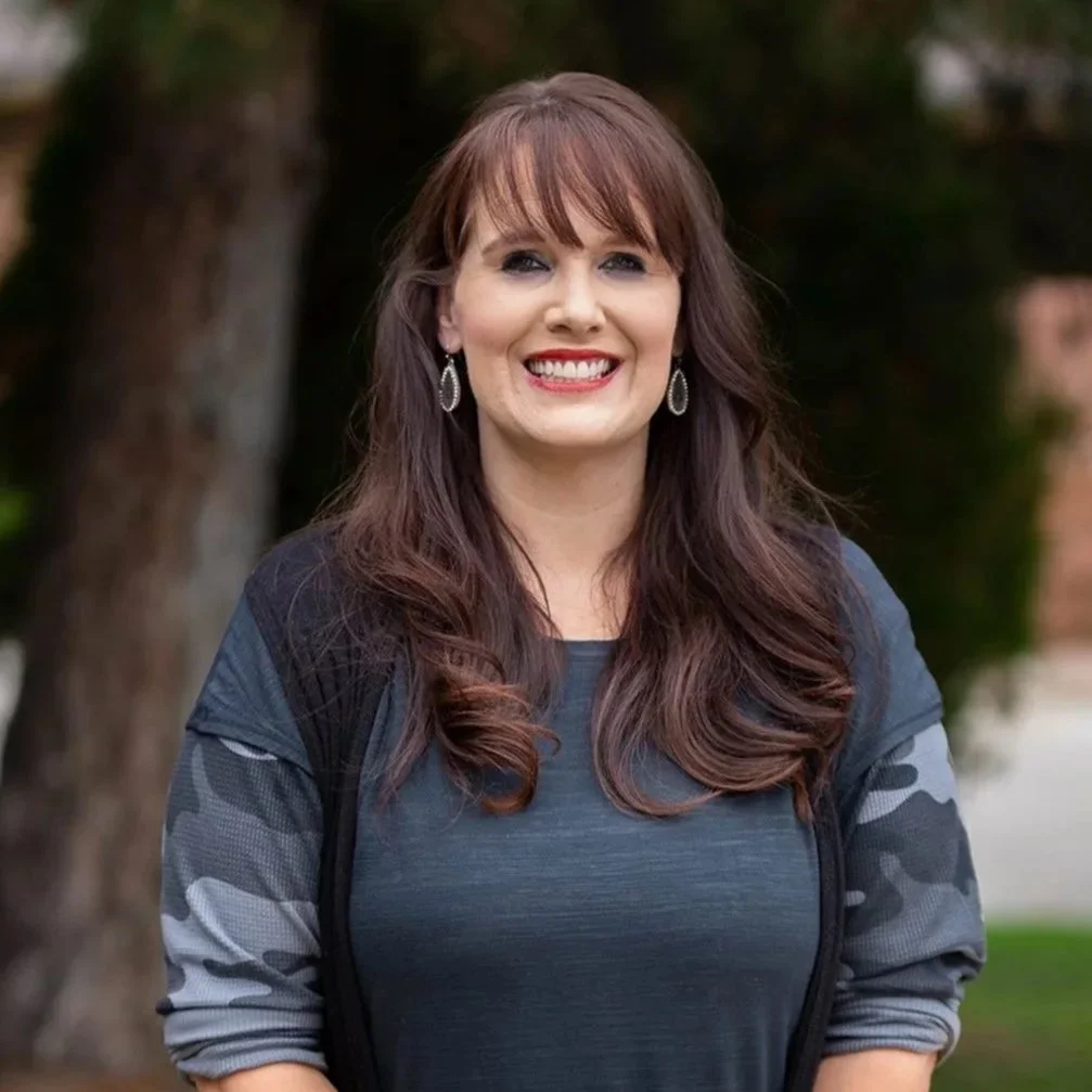 Smiling woman with long brown hair, wearing earrings, a dark gray top with camouflage sleeves, and a black vest, outdoors with trees in the background.