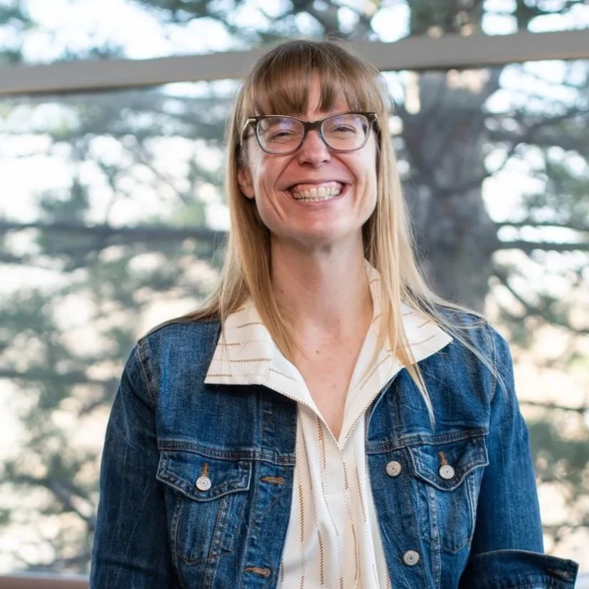 A woman with long, light brown hair and glasses smiling, wearing a denim jacket over a white, striped shirt, standing in front of a window with trees outside.