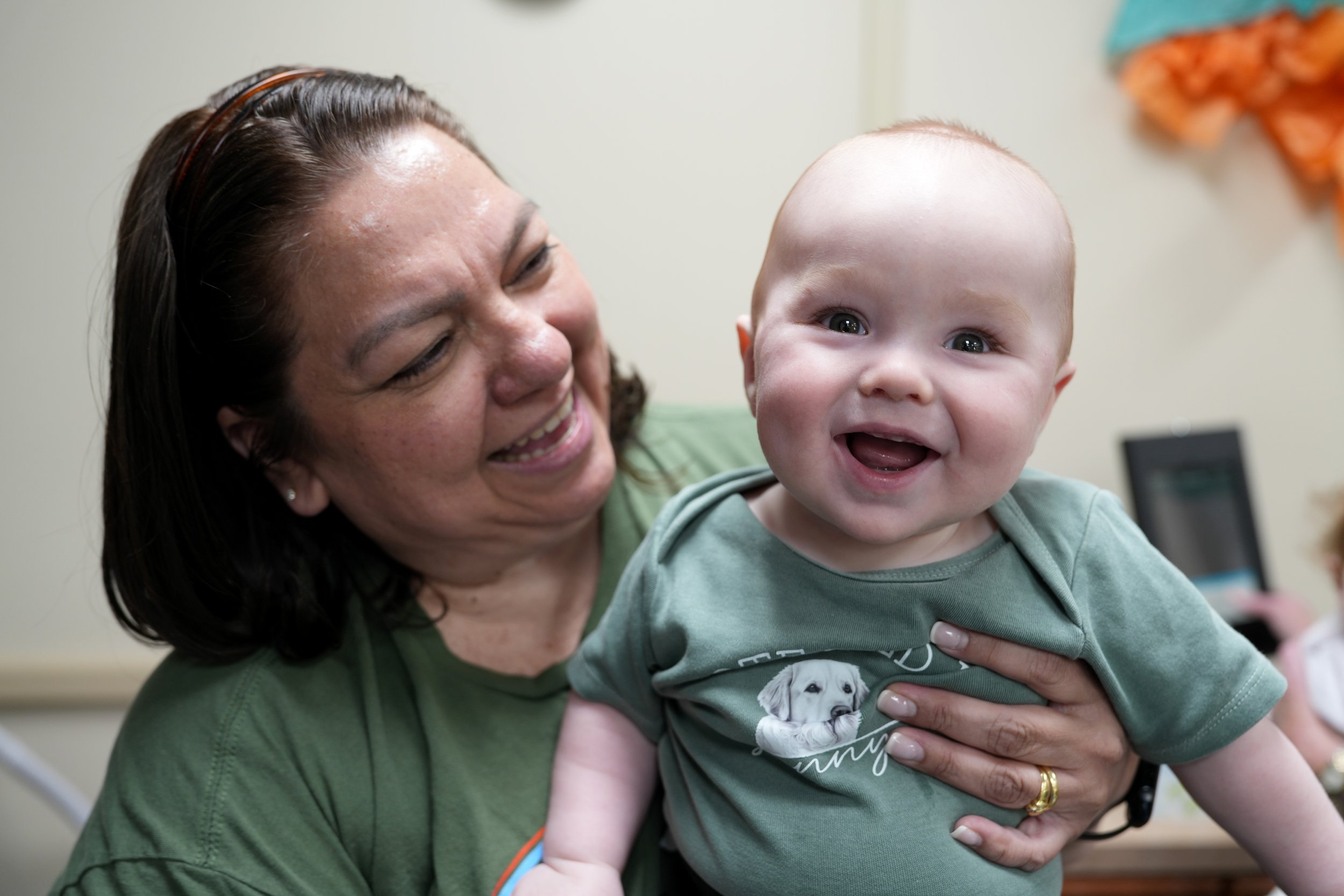 A woman with dark hair holding a smiling baby with blue eyes, both dressed casually indoors.
