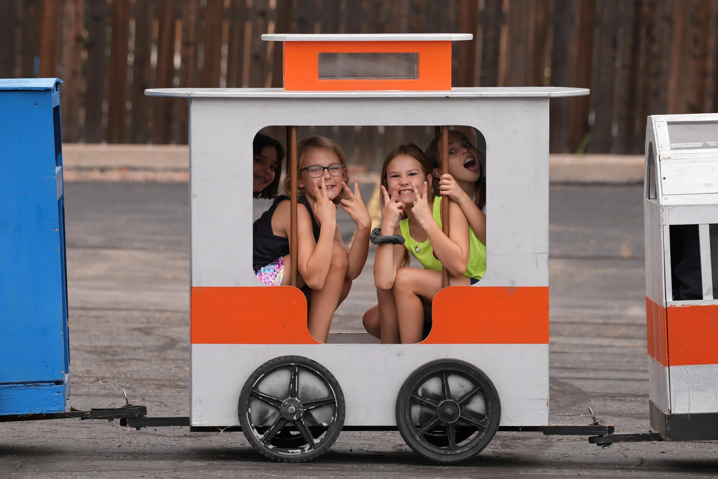 Four young girls are inside a small, orange and gray wooden toy train on wheels, pretending to drive. They are smiling and making playful expressions. The train is outdoors, with a blurred background of a wooden fence and trees.