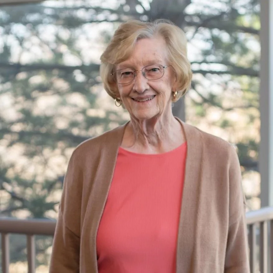 An elderly woman with short blonde hair and glasses, wearing a pink top and a tan cardigan, standing outdoors in front of a background of trees.