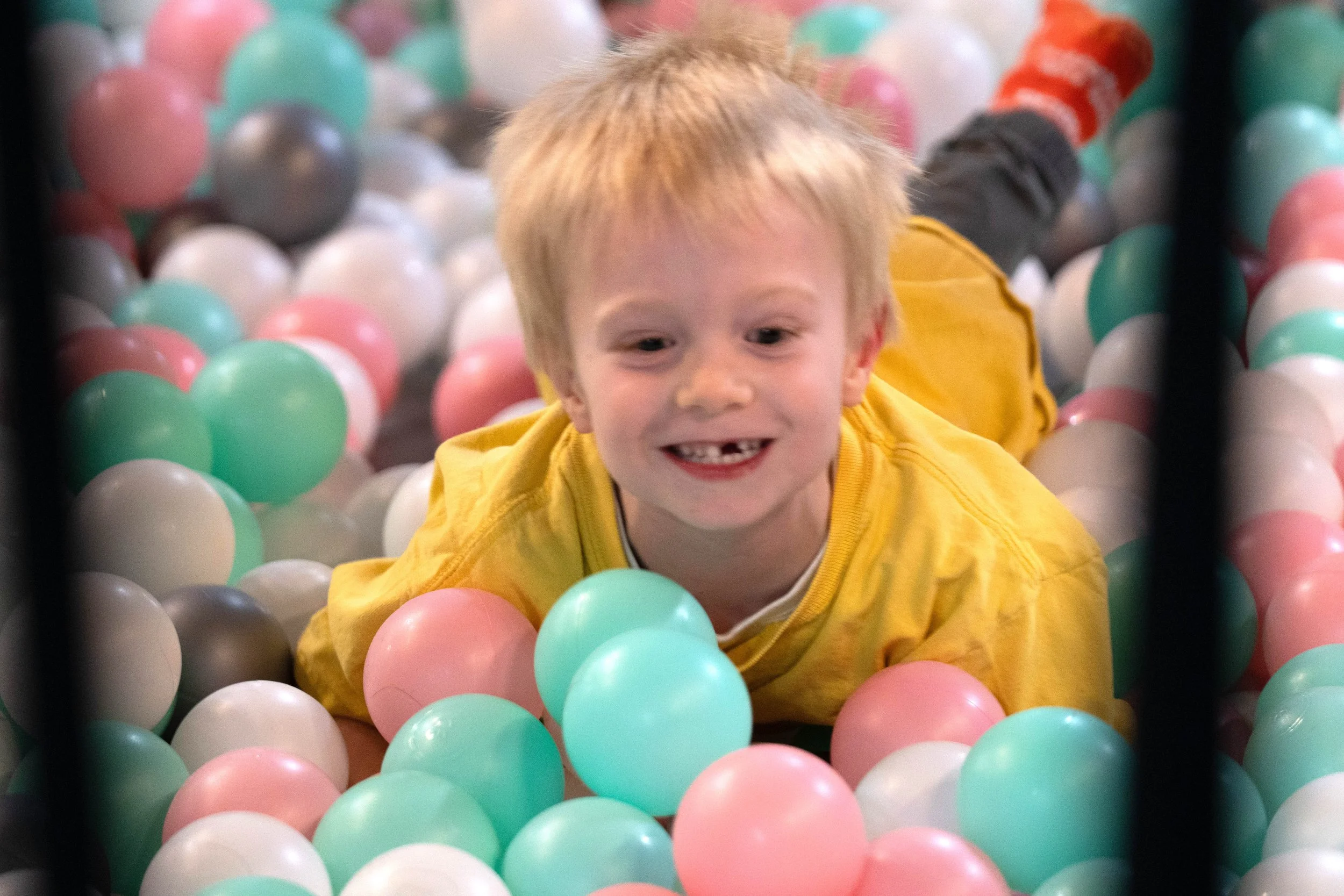 A young boy with blonde hair and a yellow shirt smiling and playing in a ball pit filled with pastel-colored plastic balls.