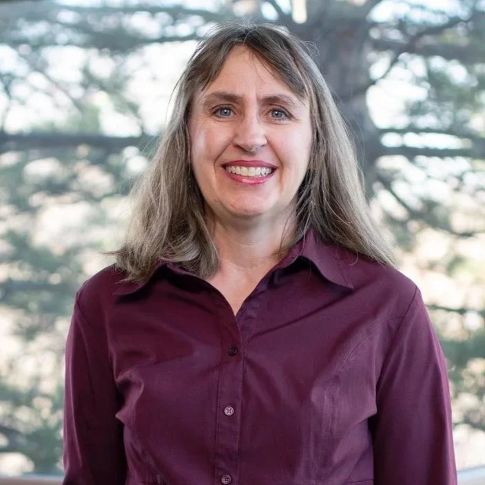 A woman with shoulder-length gray hair smiling, wearing a maroon button-up shirt, standing outdoors with pine trees in the background.