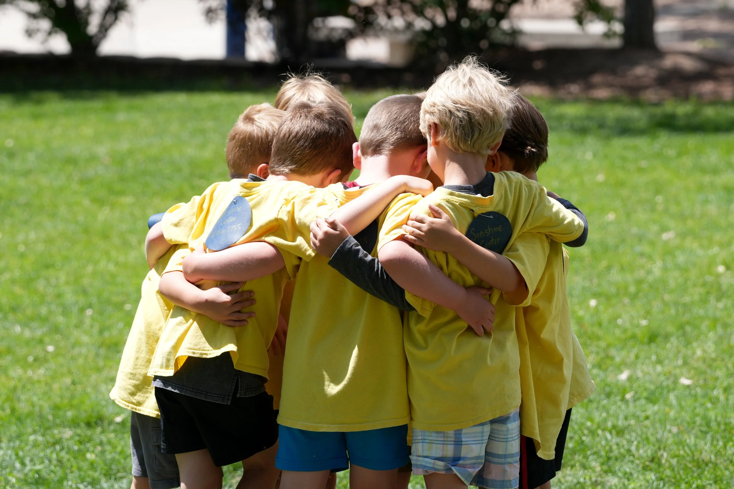 Group of children wearing yellow shirts huddled closely in a circle outdoors on a grassy area.