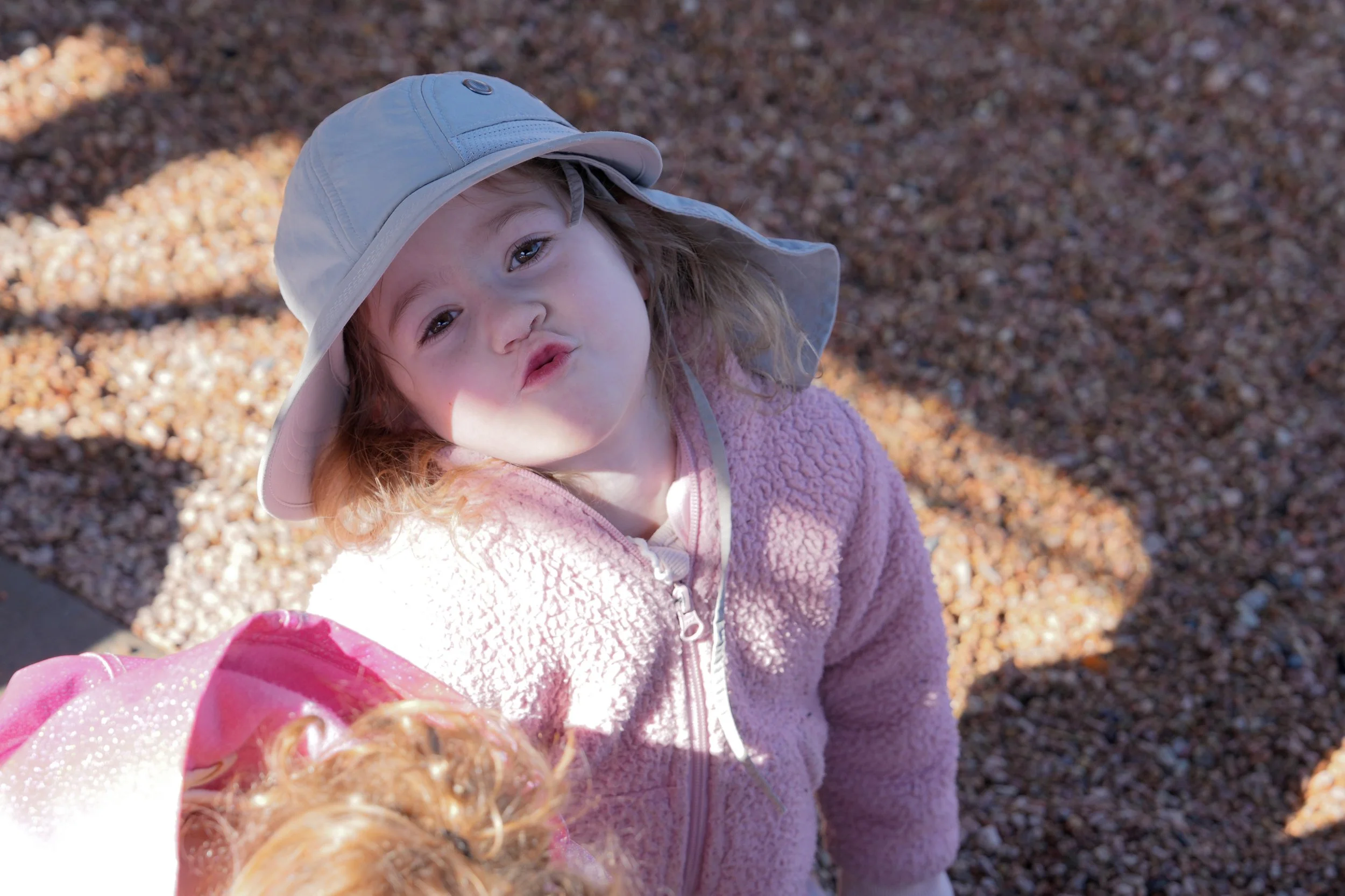 A young girl with brown hair, blue eyes, and freckles, wearing a light-colored sun hat and pink fleece jacket, looks at the camera with a kissy face. She is outside standing on ground covered with small rocks or gravel, with sunlight and shadows visible on the ground.