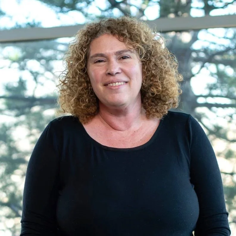 A woman with curly blonde hair smiling, standing indoors with windows and trees in the background.