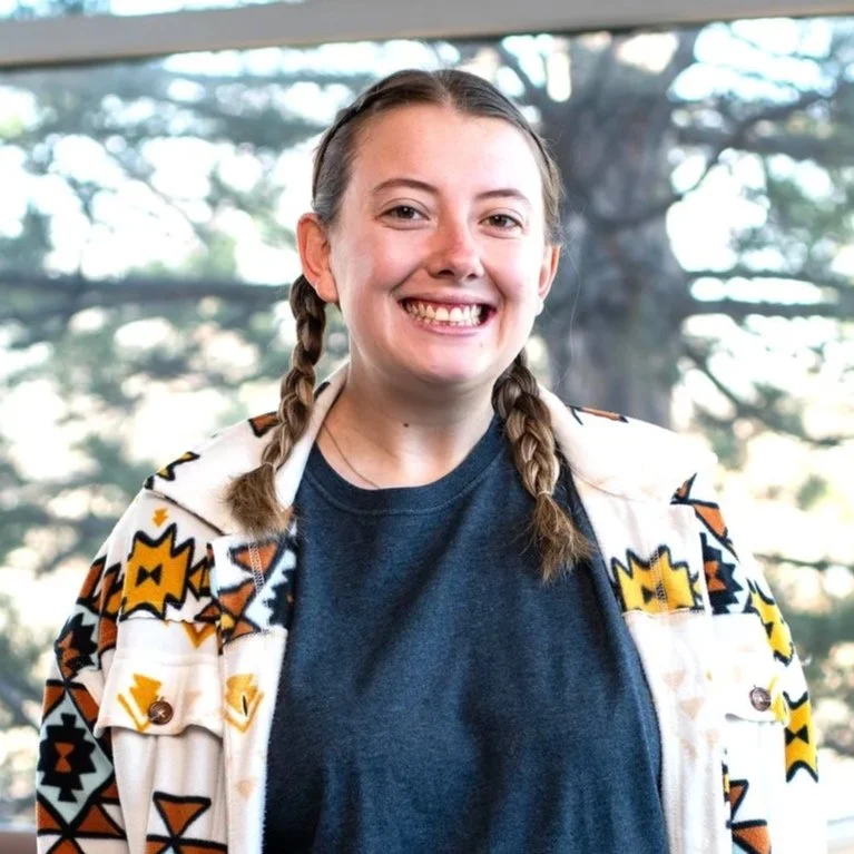 Young woman with braided hair smiling, wearing a patterned jacket and a dark t-shirt, standing in front of a window with trees outside.