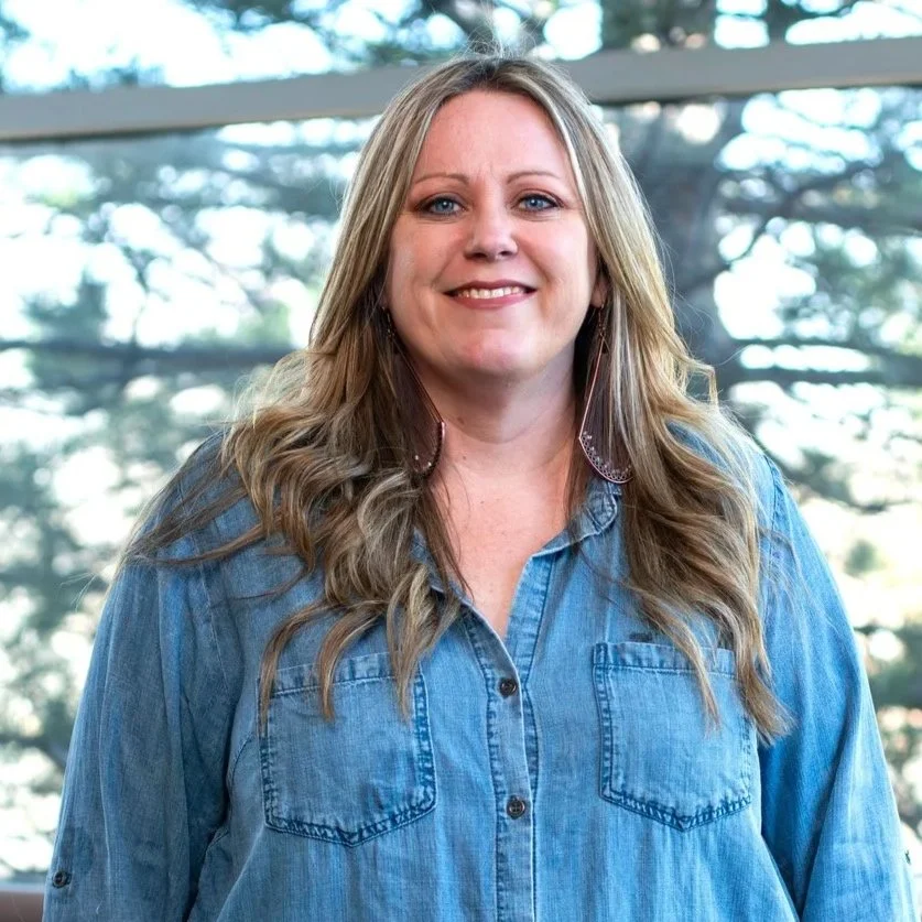 A woman with long blonde hair smiling, wearing a denim shirt, standing indoors with a window showing trees outside.