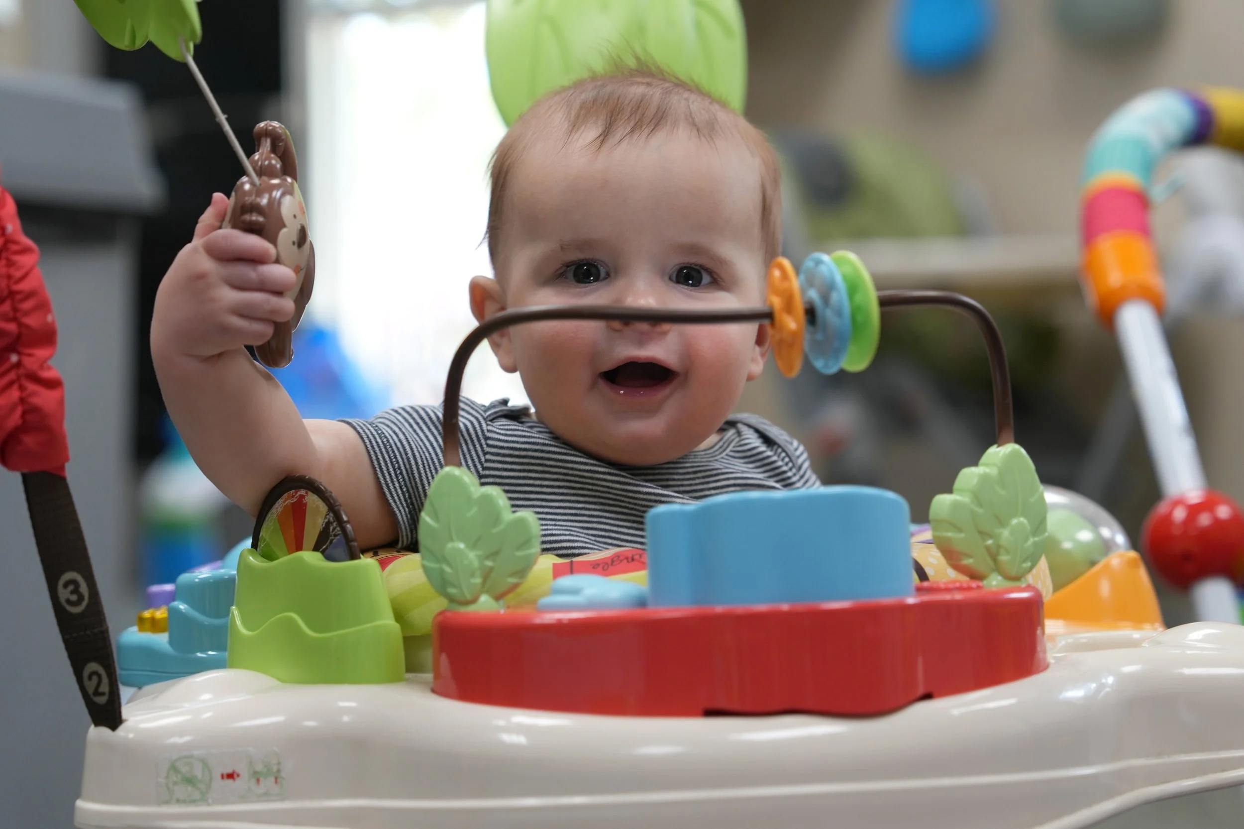 A young child sitting in a colorful baby walker, holding a toy in one hand, surrounded by various toys and colorful elements.