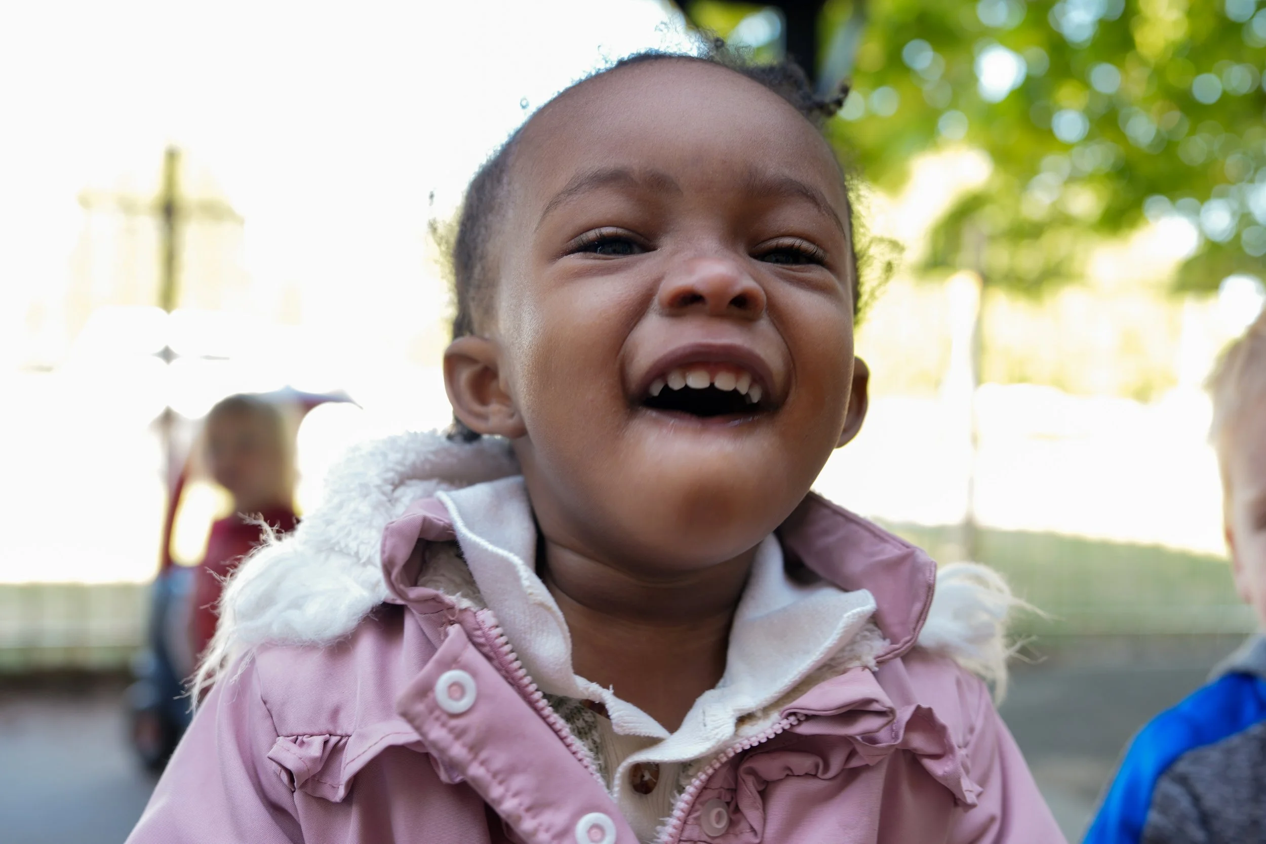 Close-up of a smiling young girl wearing a pink jacket outdoors with greenery and blurred children in the background.