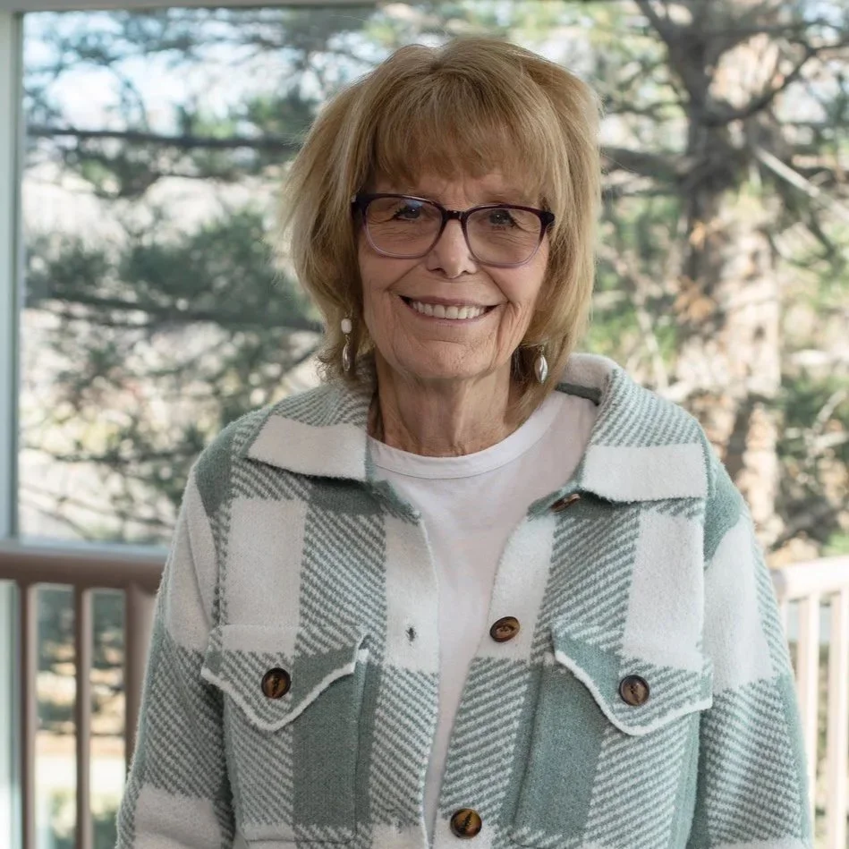 An elderly woman with glasses smiling outdoors, wearing a plaid jacket and a white shirt, with trees in the background.