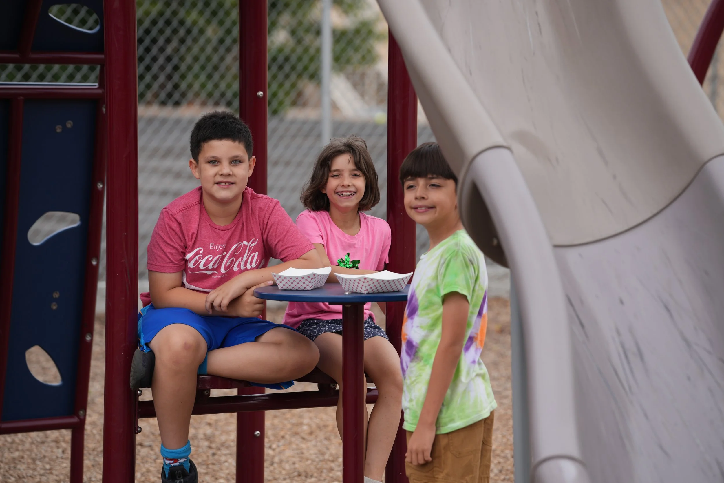 Three kids, two boys and one girl, sitting at a table on playground equipment, smiling. Another boy, wearing a tie-dye shirt, stands nearby. It is outdoors, with a slide and chain-link fence in the background.