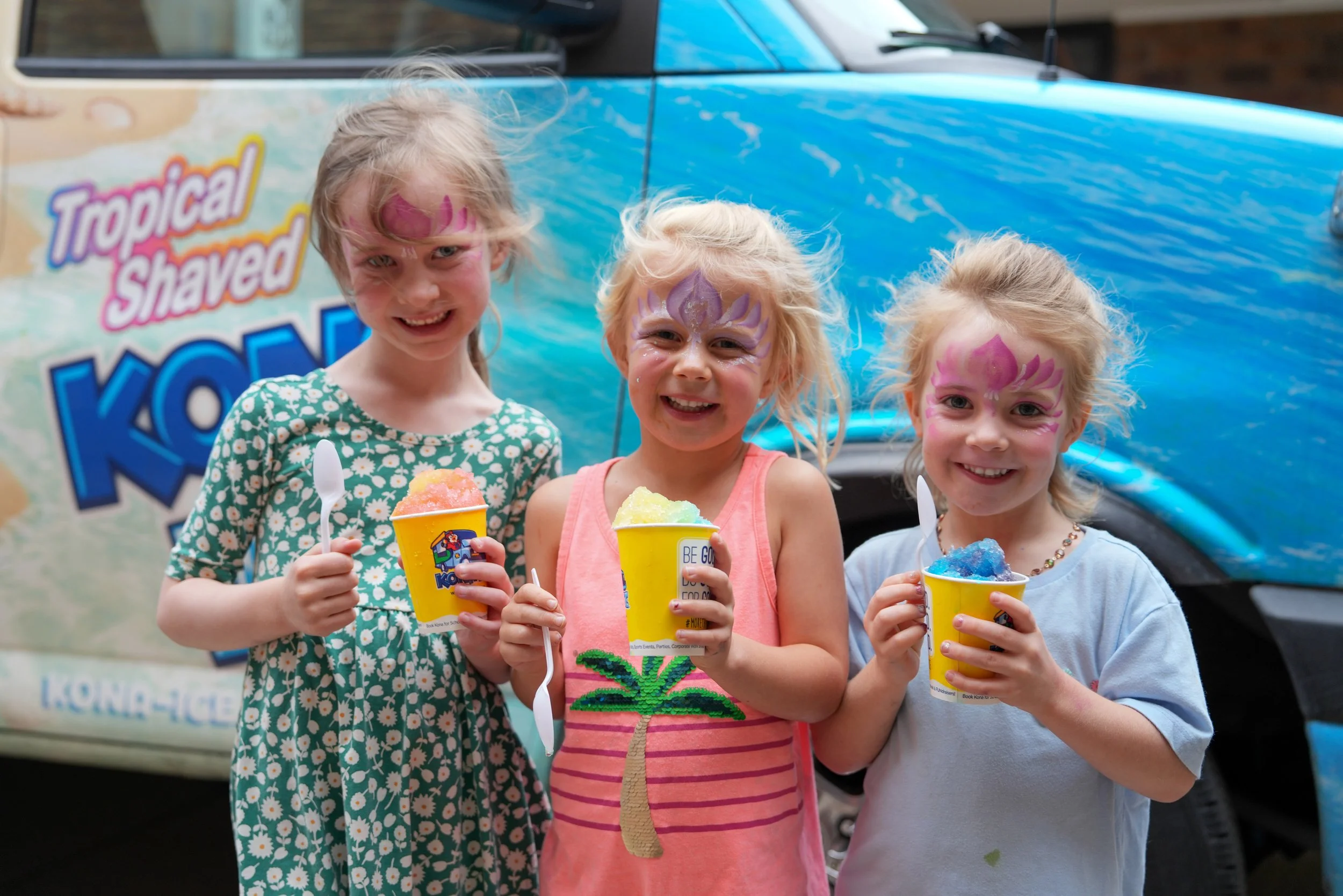 Three young girls with face paint holding colorful cups of snow cones, standing in front of a blue vehicle with a large advertisement promoting tropical shaved ice.