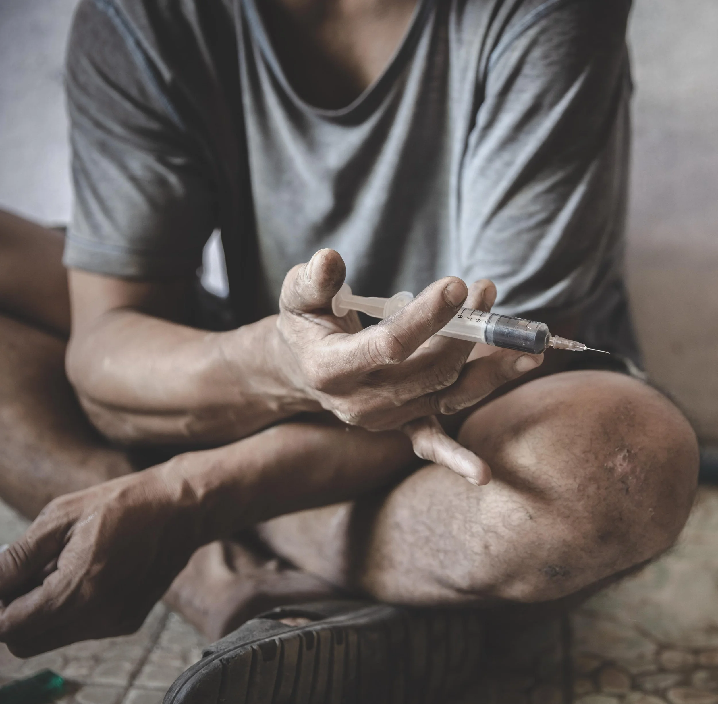 A person sitting cross-legged appears to be receiving an injection from another individual, with a focus on the syringe in their hand.