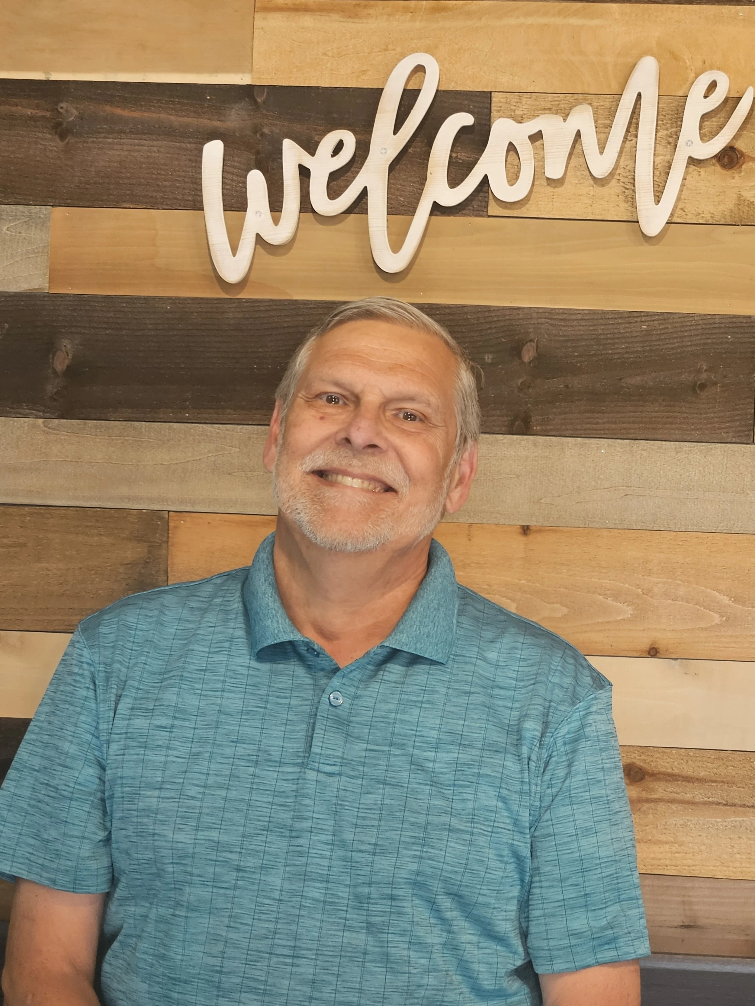 A man with gray hair and a beard smiling in front of a wooden wall with a sign that says 'welcome'.