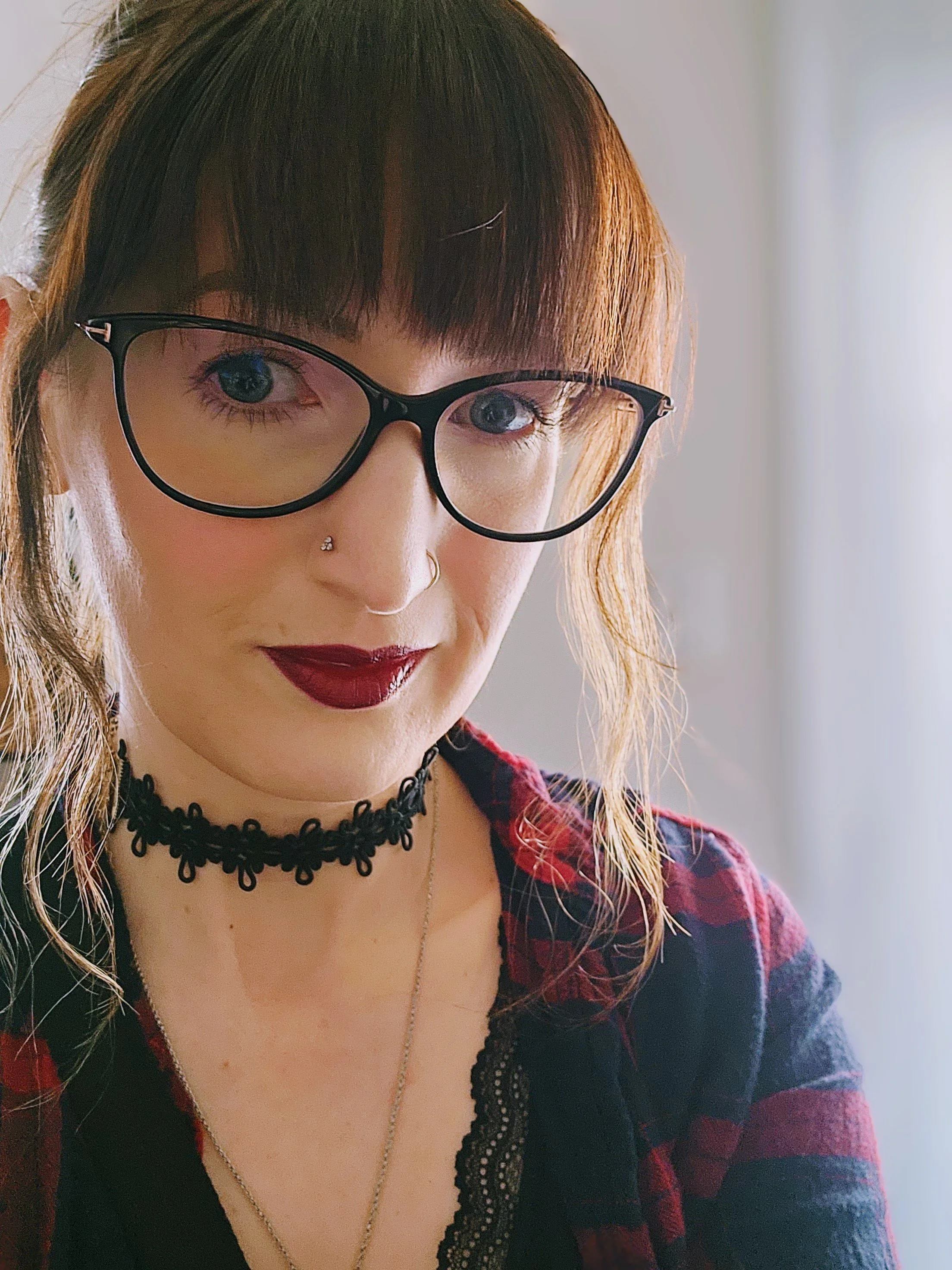 A woman with light skin and dark brown hair, wearing black-rimmed glasses, a black lace top, layered necklaces, a black choker with a floral design, and a red and black plaid shirt, posed indoors near a window with soft lighting.