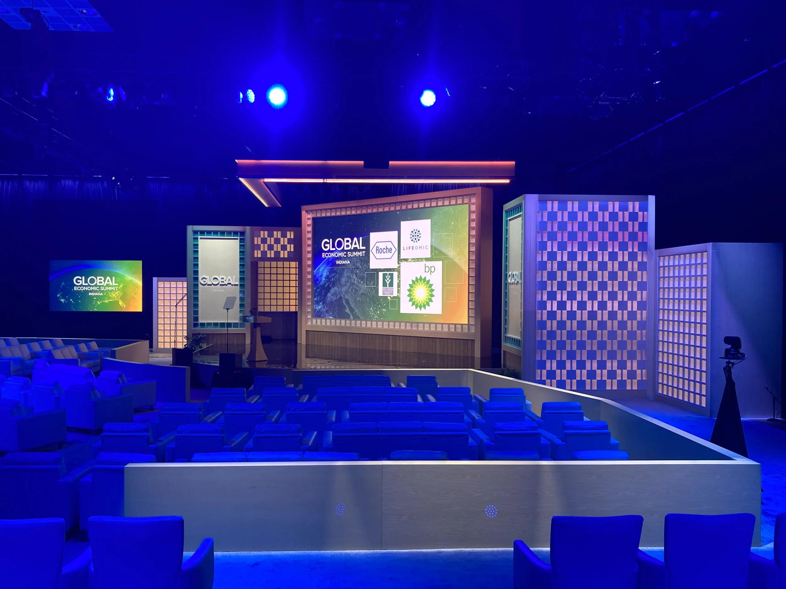 Conference stage setup with large screen displaying logos, blue lighting, and rows of empty chairs.