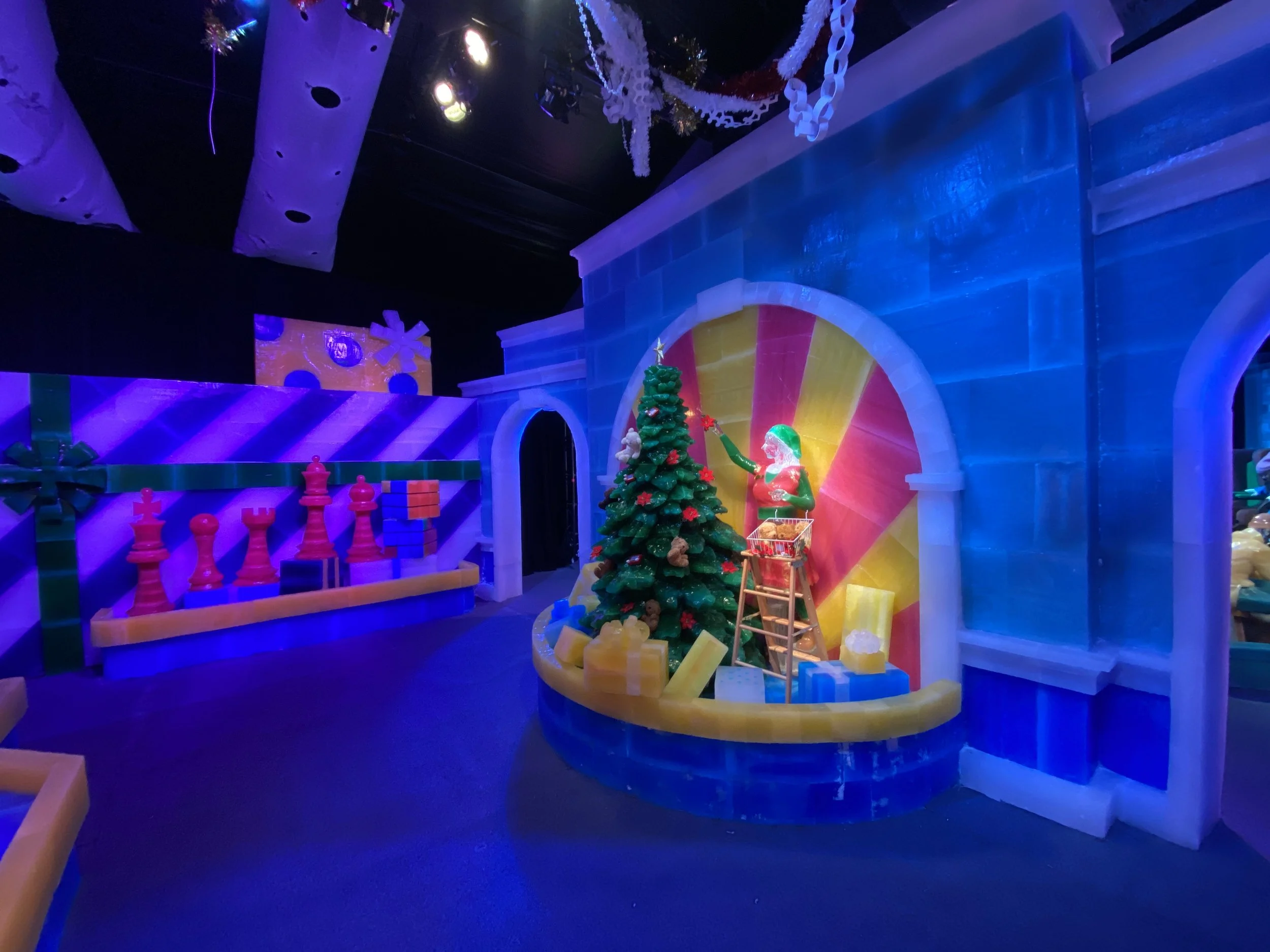A brightly lit indoor space decorated for Christmas, featuring a Christmas tree, a festive backdrop with wrapped presents, and various holiday-themed decorations, illuminated with colorful lighting.