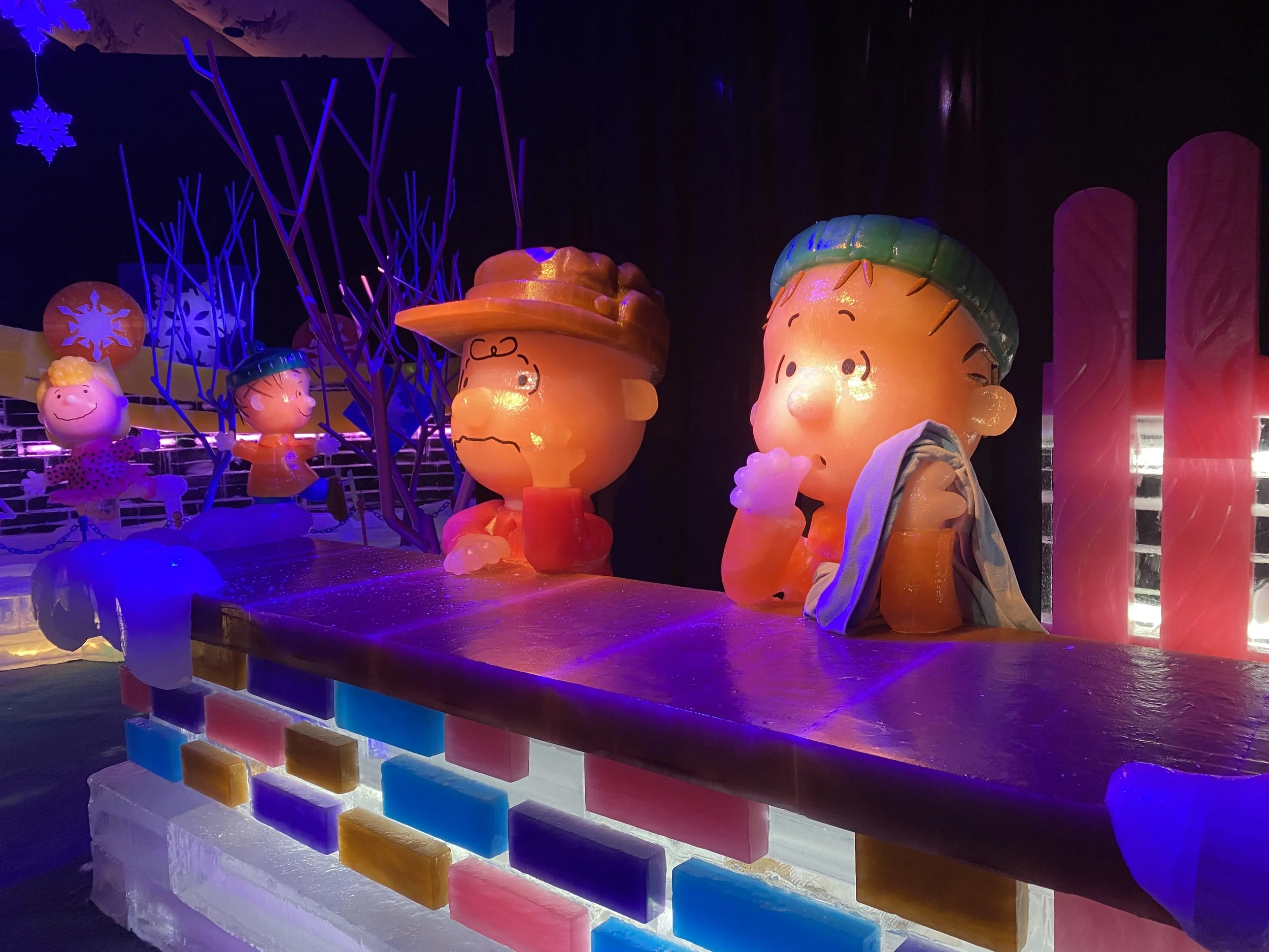 Colorful ice sculptures of Charlie Brown, Sally, and other Peanuts characters, illuminated in a dark room with purple and pink lighting.