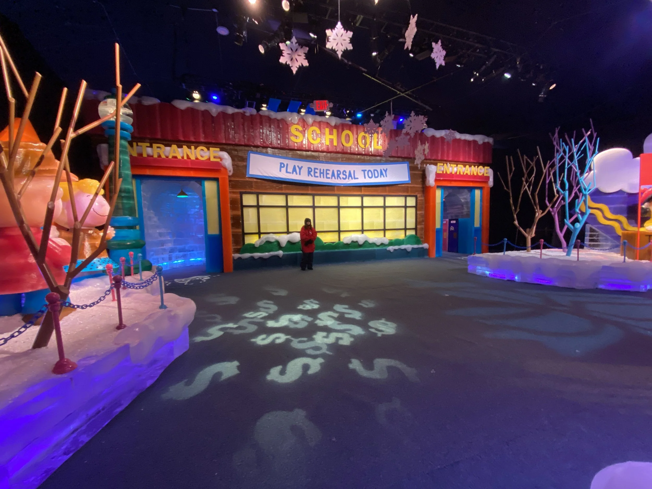 Indoor ice skating rink decorated for winter with snowflake decorations, colorful lights, and a festive sign that reads 'Play Rehearsal Today'.