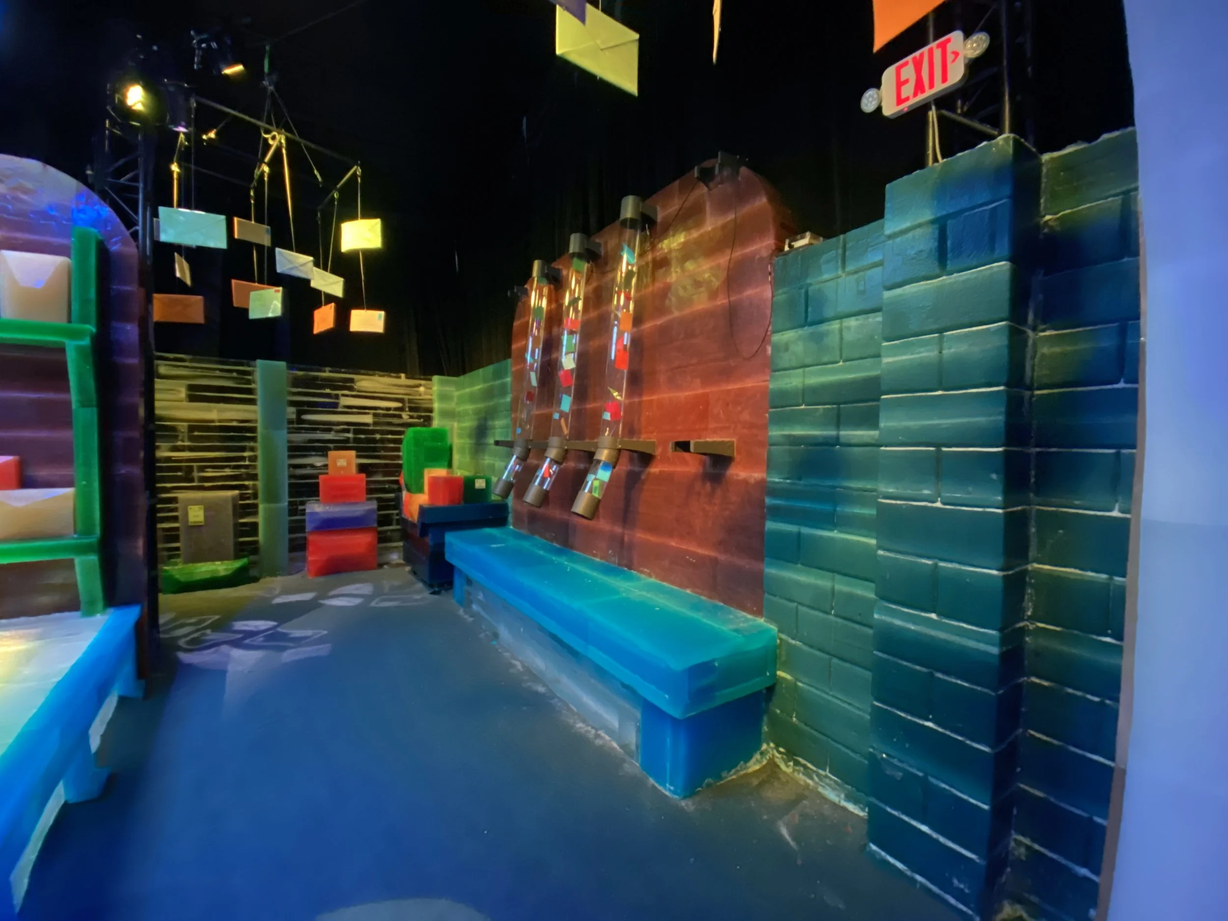 Colorful indoor playground with padded walls, climbing structures, slides, and hanging rectangular flags or banners.