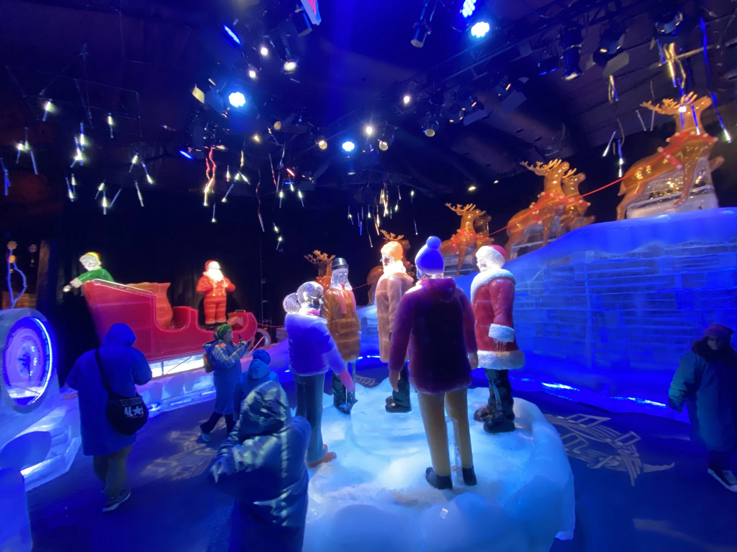 People walking inside a dimly lit ice skating or winter-themed attraction with holiday decorations, including reindeer and Santa figures, illuminated by blue and white lights.