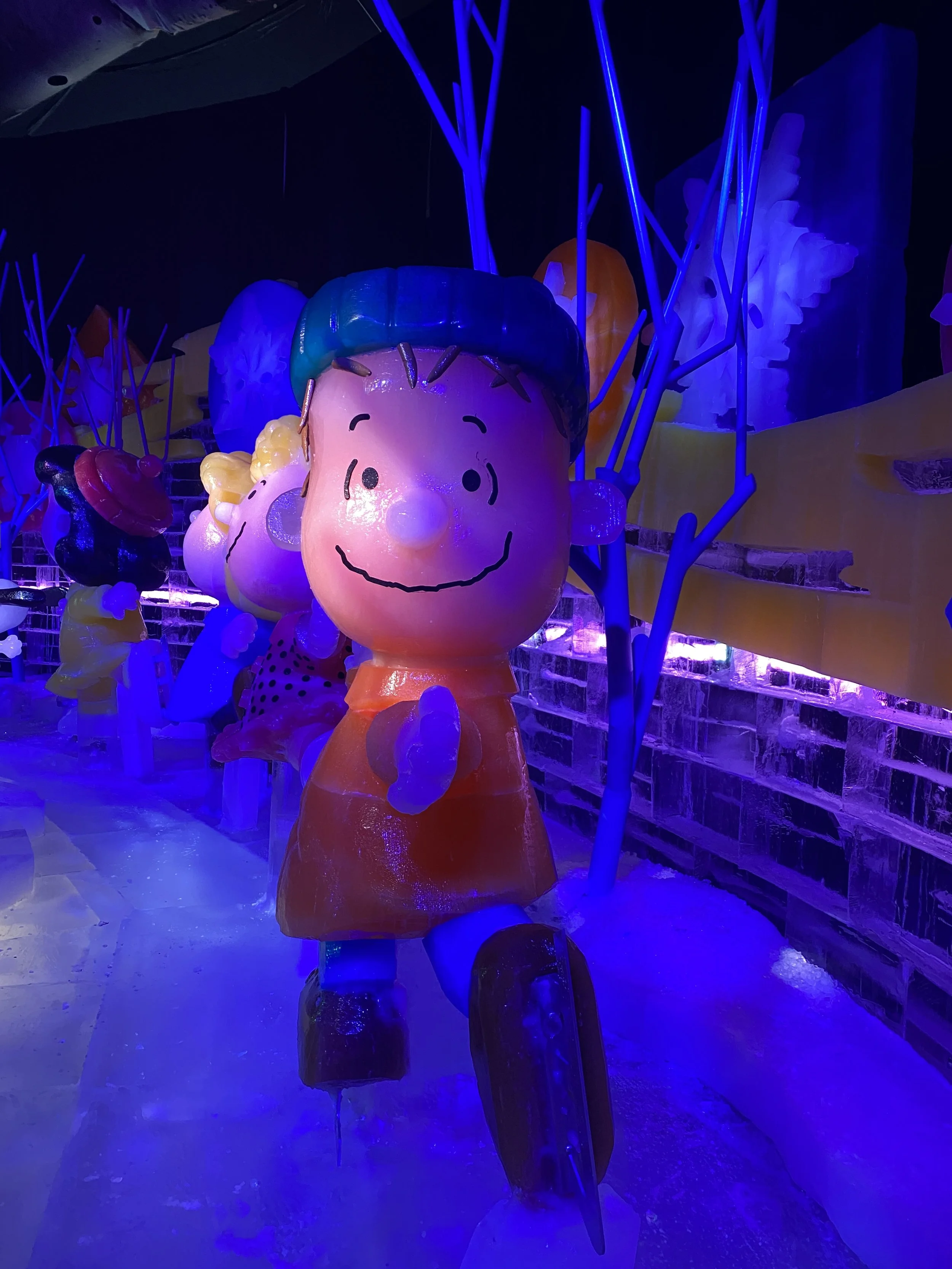 Colorful ice sculpture of Charlie Brown with blue hair, surrounded by other ice figures and decorated with neon blue lights.