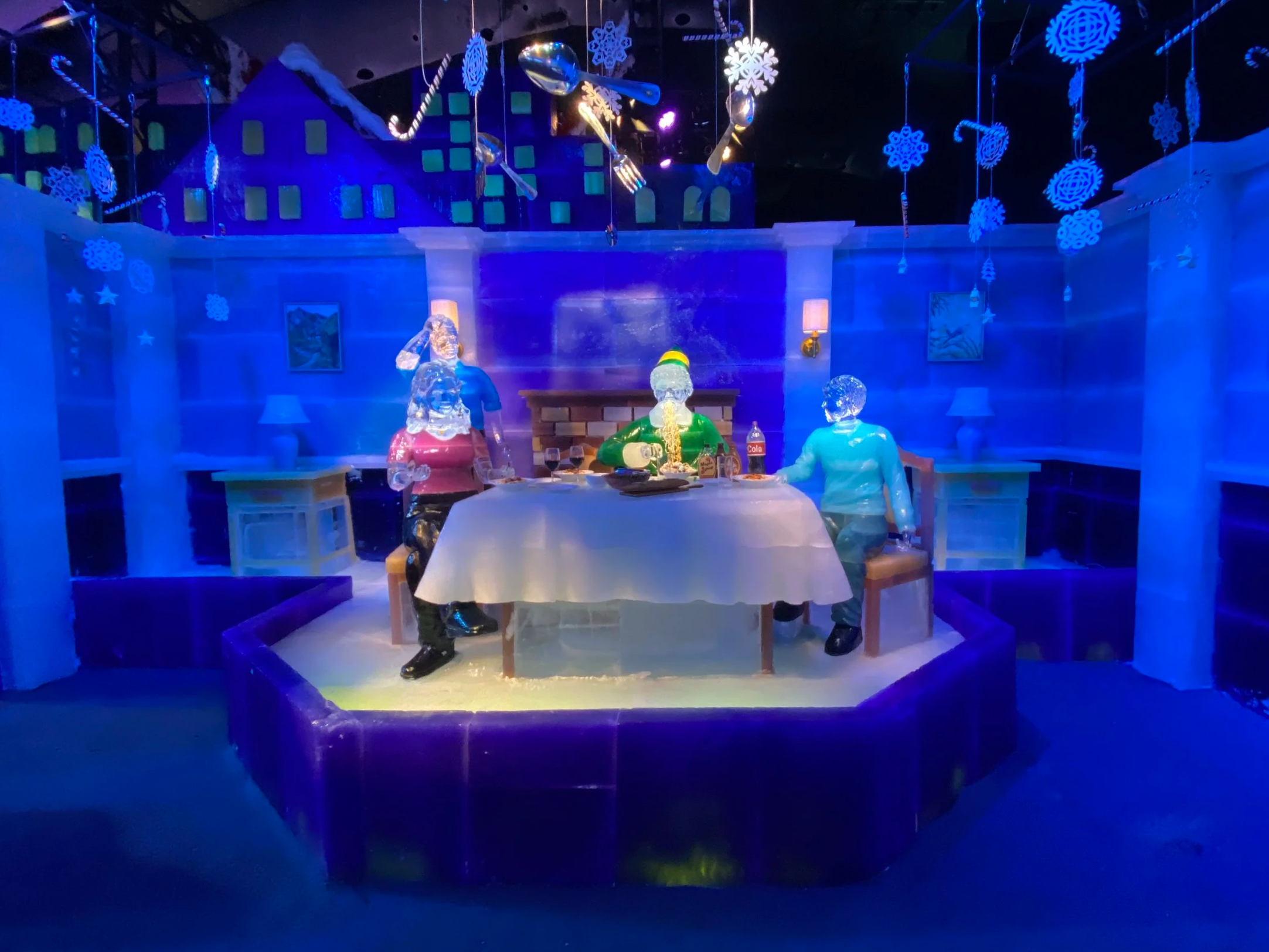 A holiday-themed ice sculpture display with three figures sitting at a table, surrounded by hanging snowflake decorations and illuminated with blue and purple lighting.